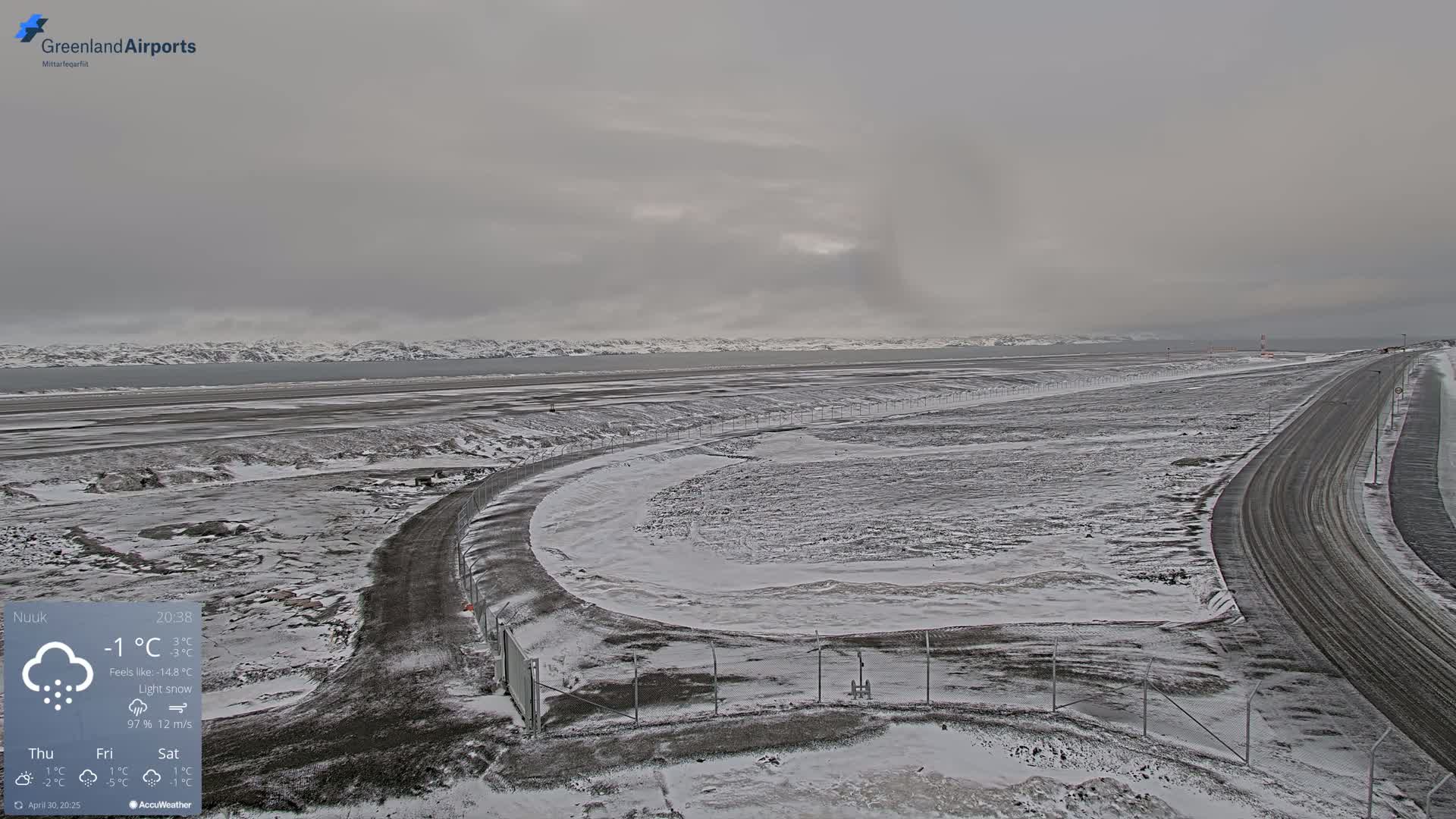 A snow-covered airport with a road curving through the tarmac, under a light snow in overcast conditions, overlooks a body of water and distant mountains.