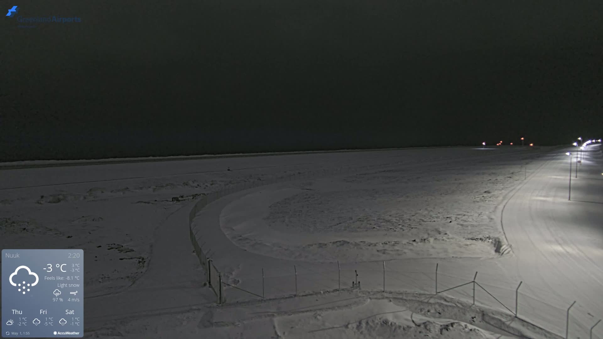 A snow-covered, dimly lit airport runway and surrounding area are seen at night under light snow.