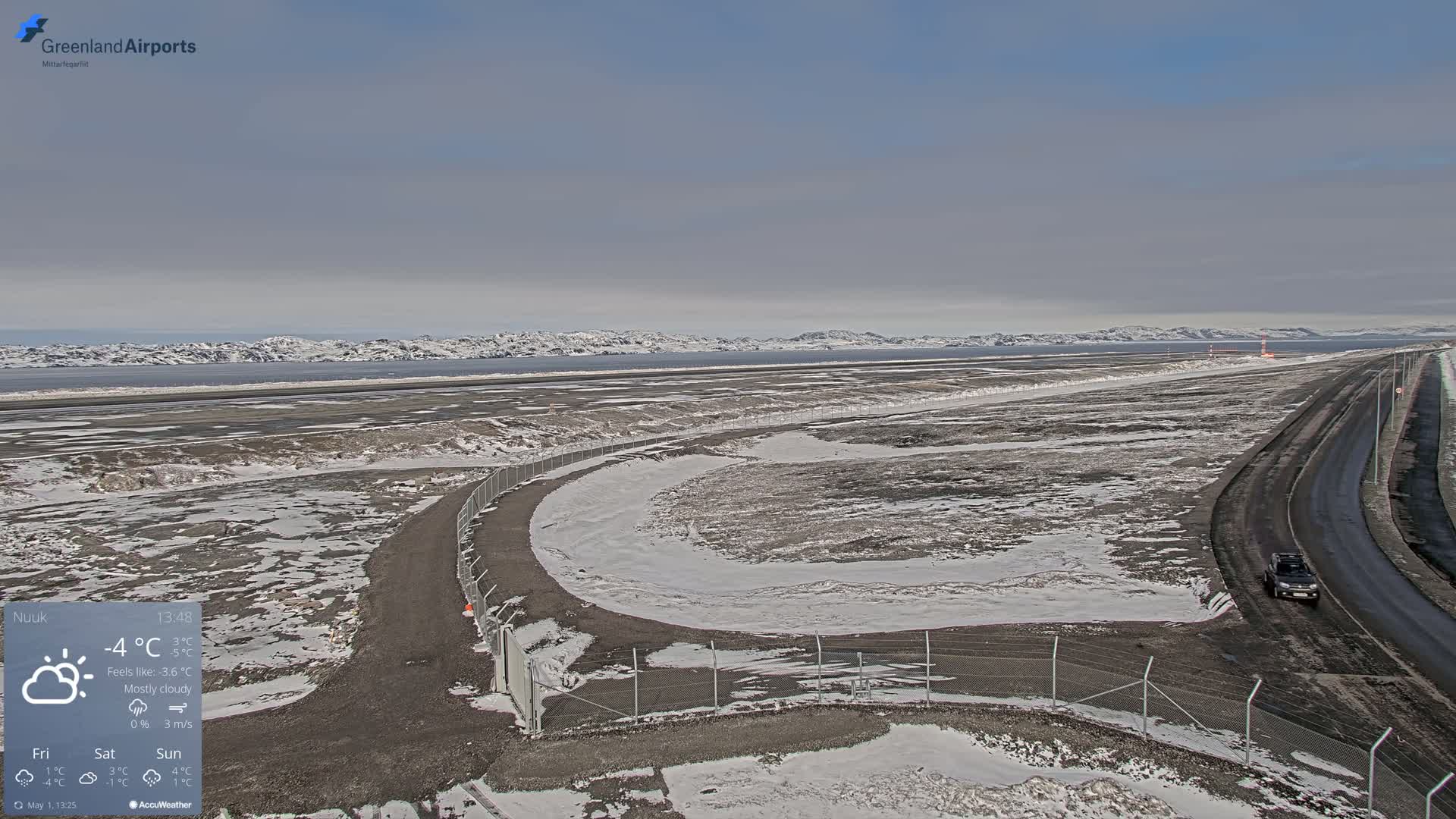 A mostly cloudy, snowy airfield with a road and a vehicle is seen, with mountains and a body of water visible in the distance.