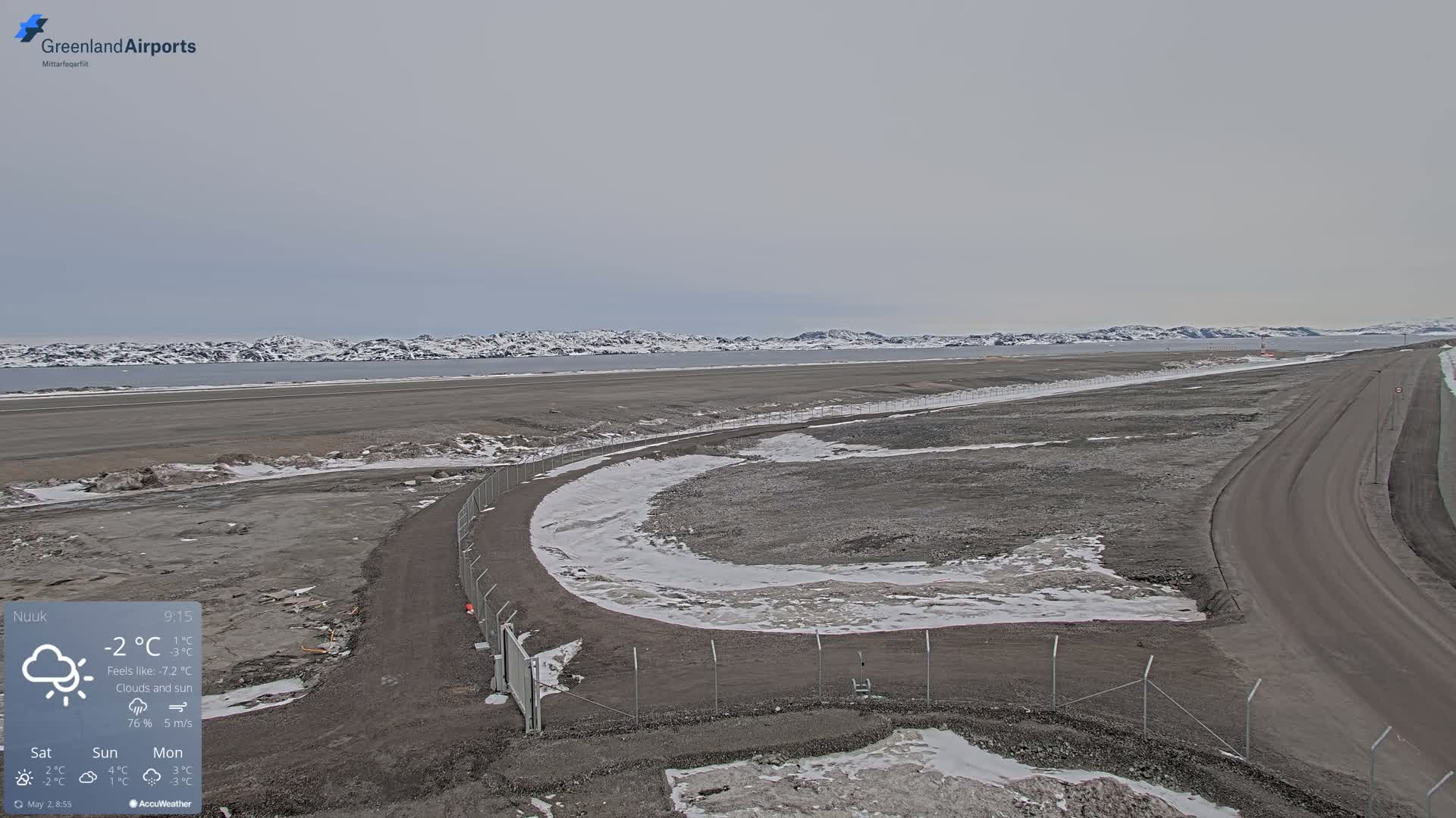 A mostly cloudy and snowy airfield with a road and fence leading to a body of water, backed by snow-covered mountains.