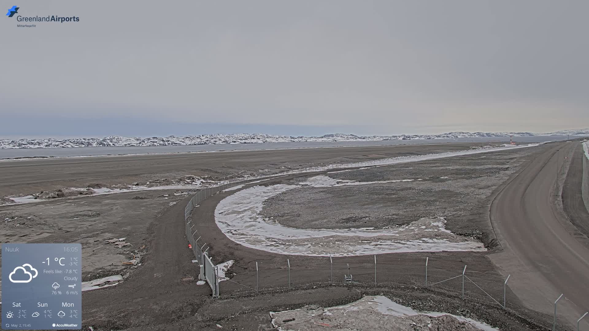 A mostly cloudy, cold day reveals a view of a flat, gray landscape with a road curving near a chain-link fence, a body of water, and snow-capped mountains in the distance.