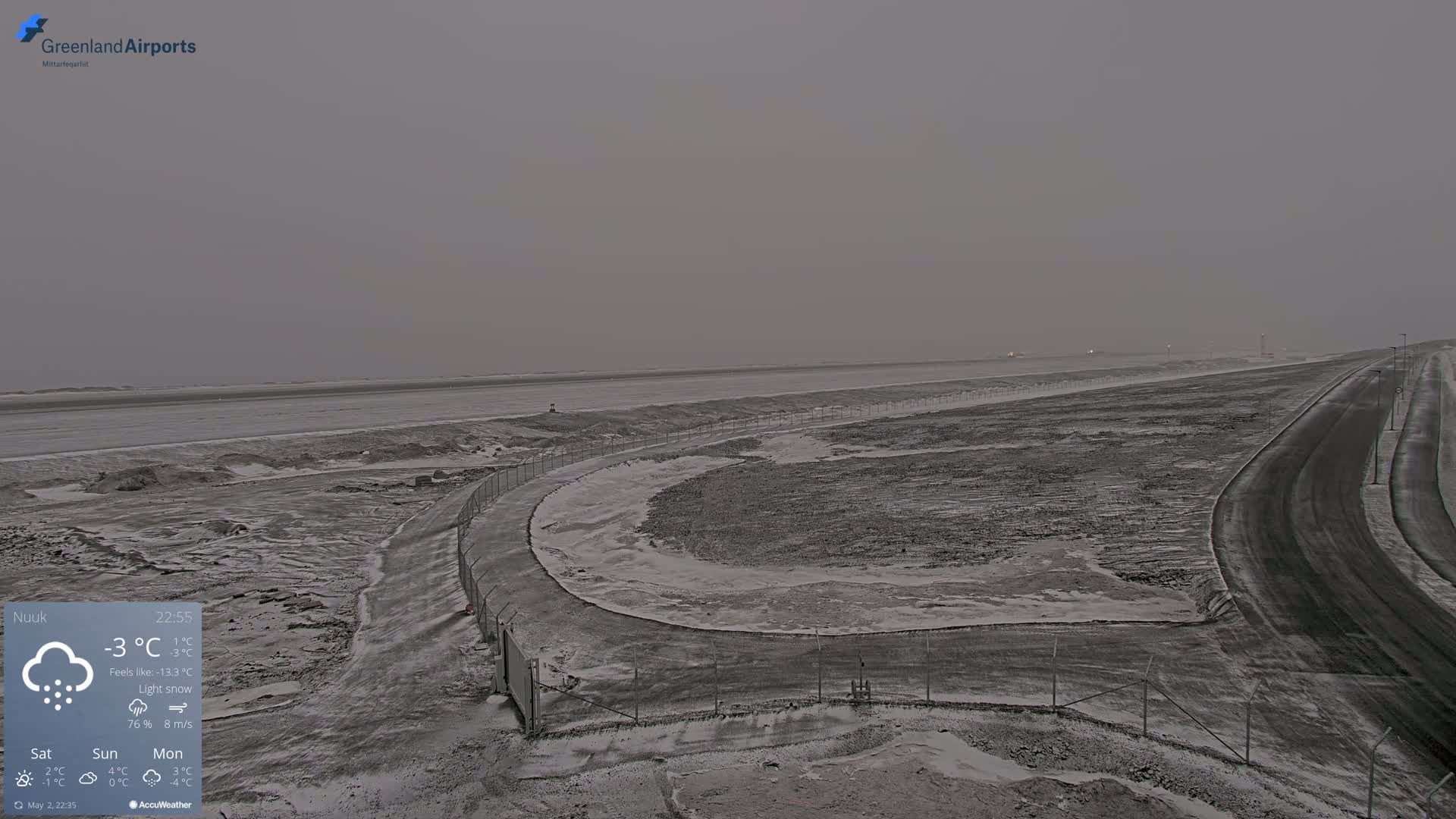 A snow-covered landscape with a road curving through it under a gray, overcast sky, light snow is falling.