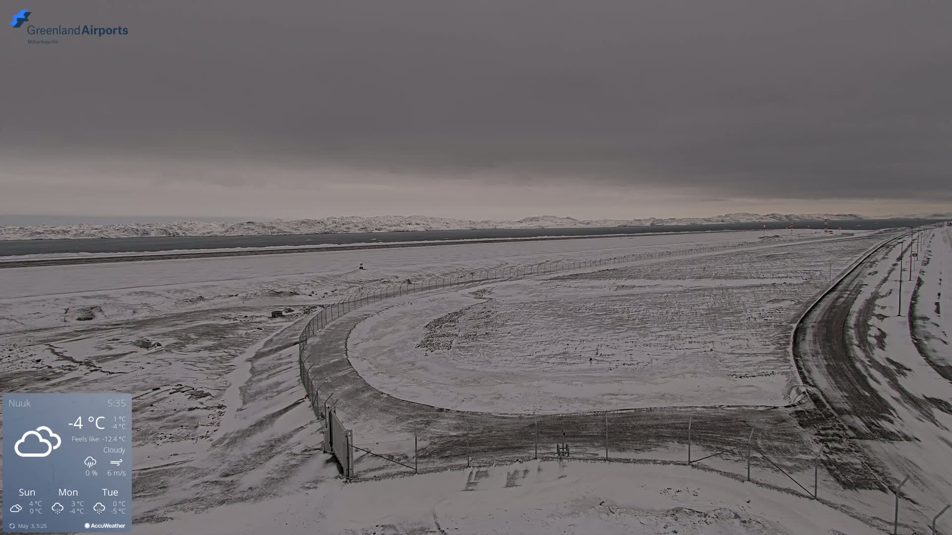 A snow-covered airport runway and road extend toward a distant, snow-capped mountain range under a cloudy sky.