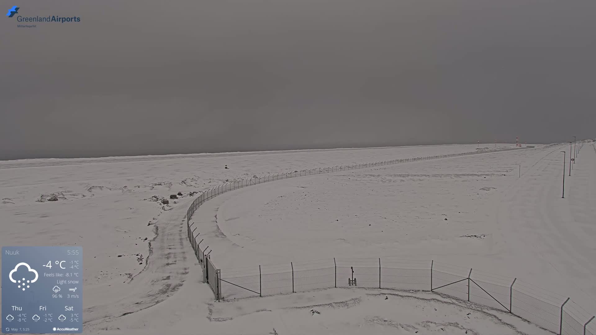 A snow-covered landscape with a chain-link fence and a road under a gray, overcast sky with light snow.