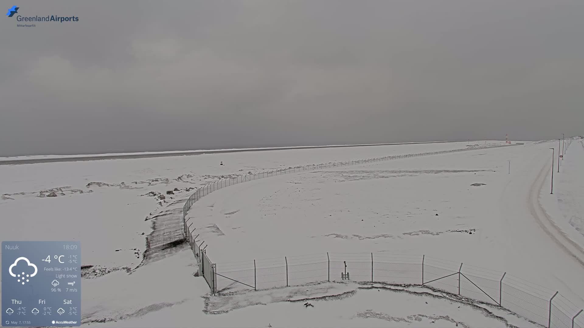 A snow-covered landscape with a chain-link fence curving around a section of land under a light snow in overcast conditions.