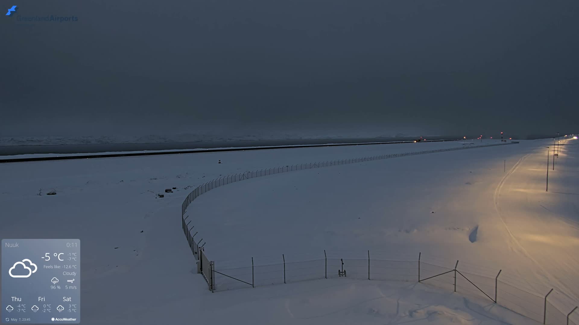 A snow-covered landscape at twilight, featuring a fence, a road with lights, and a body of water in the distance under cloudy skies.