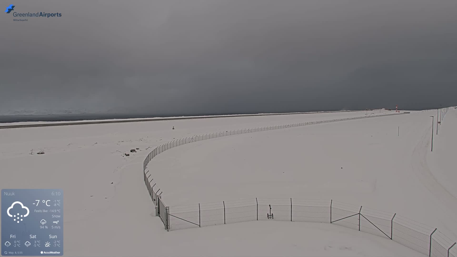 A snow-covered airfield is visible under an overcast sky, with a chain-link fence curving through the foreground.