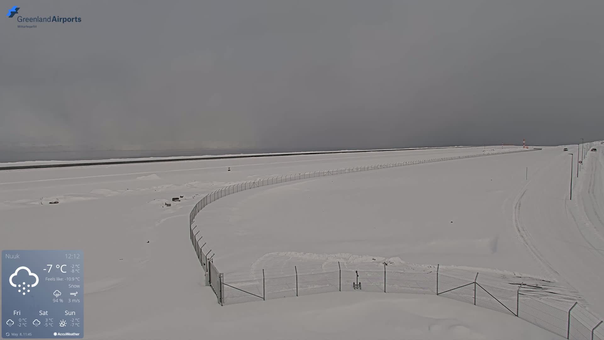 A snow-covered airfield with a chain-link fence is seen under a gray, overcast sky, snowing lightly.