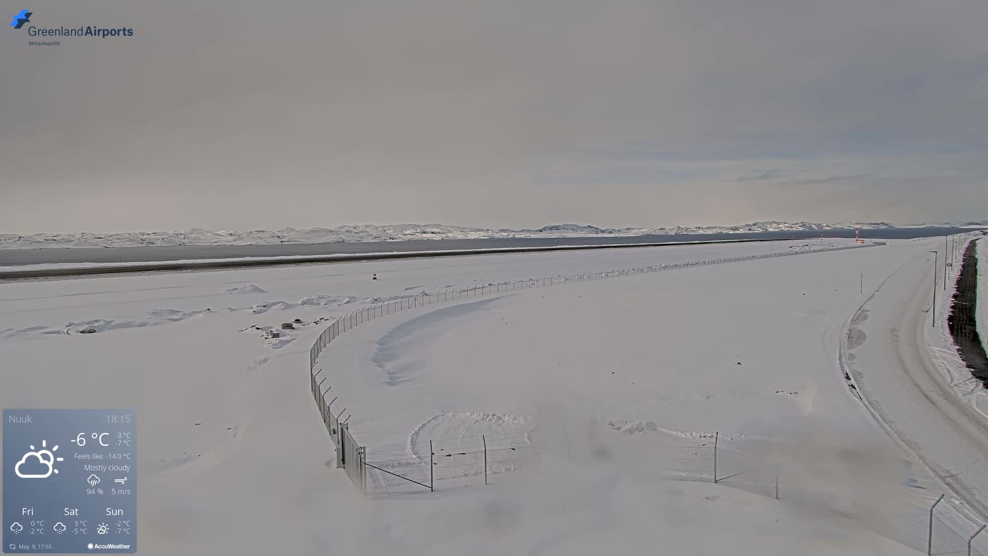 A snow-covered airport runway and road extend toward a distant, snow-capped mountain range under a mostly cloudy sky.