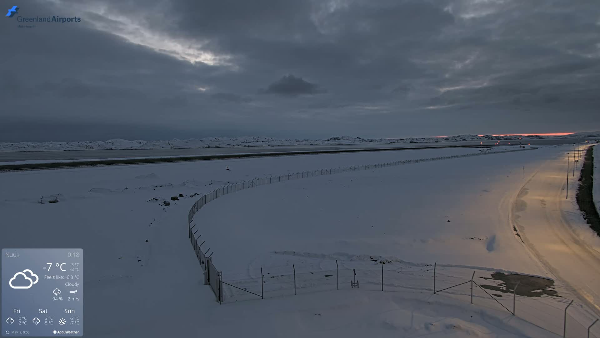 A snow-covered landscape at dusk, featuring a road, a fence, and a body of water in the distance under a cloudy sky.