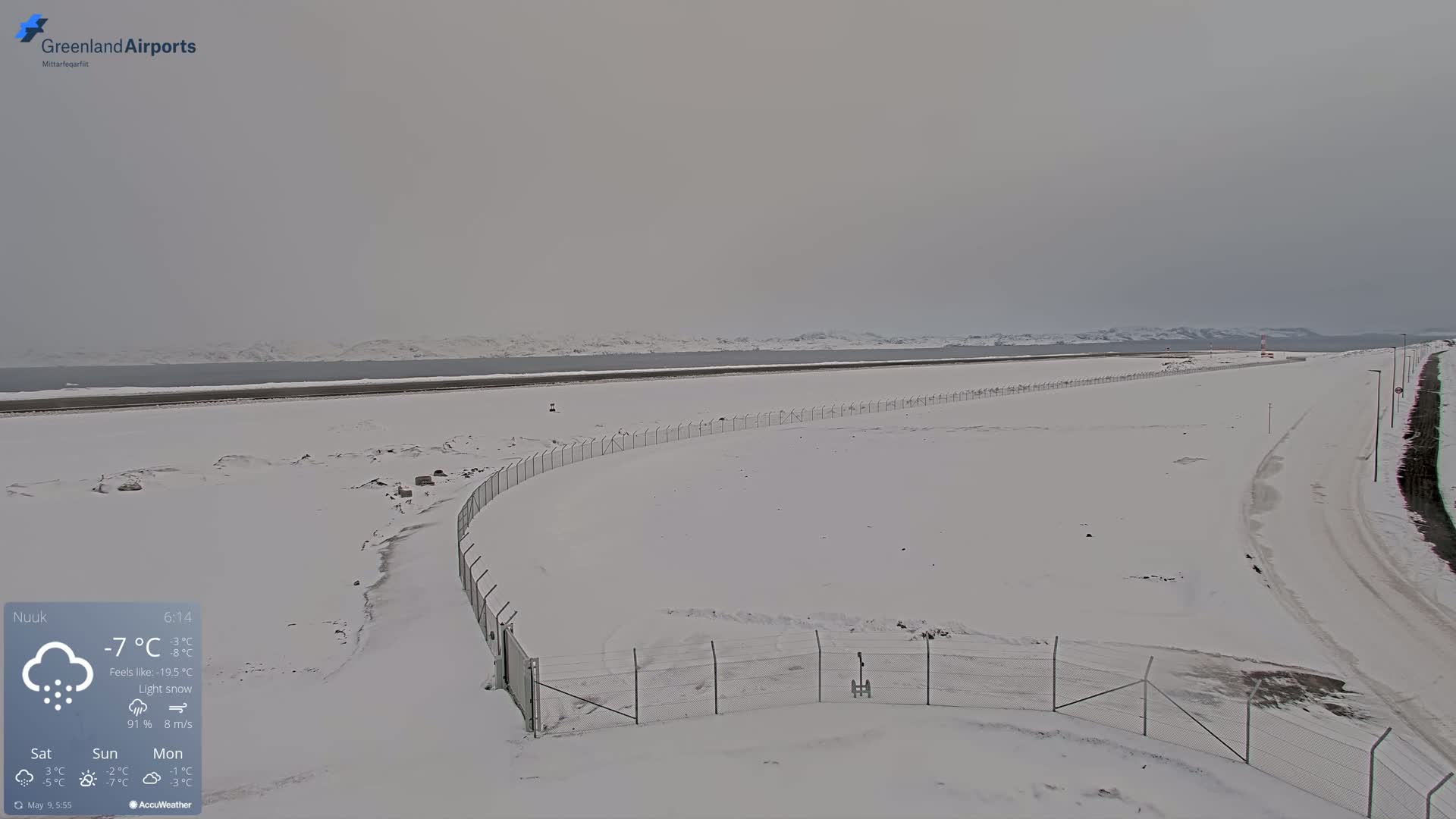 A snow-covered airfield with a chain-link fence stretches toward a calm, gray body of water under an overcast sky.