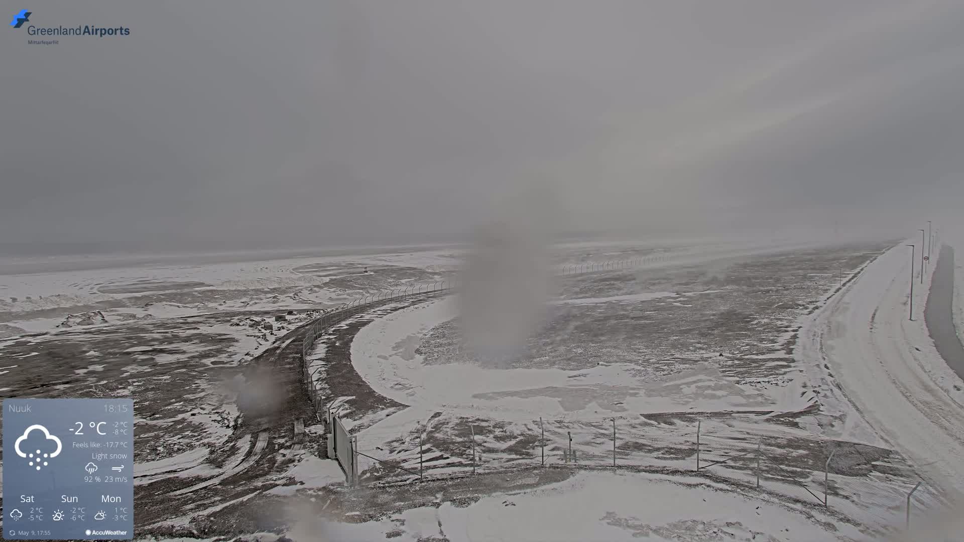 A snow-covered landscape with a road and fence is visible under a light snow, with blowing snow obscuring the distance.