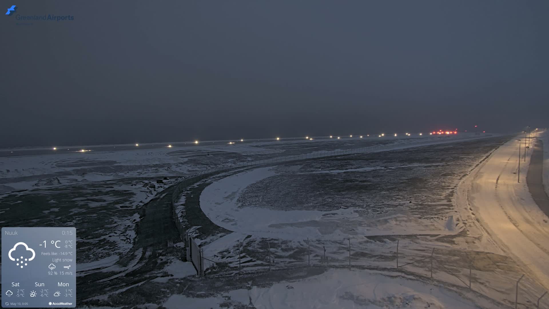 A snow-covered airport runway and road are illuminated at night under light snow.