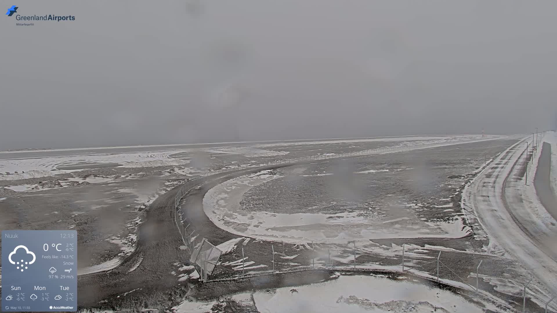 A snow-covered landscape with a road curving near a body of partially frozen water under a gray, overcast sky.