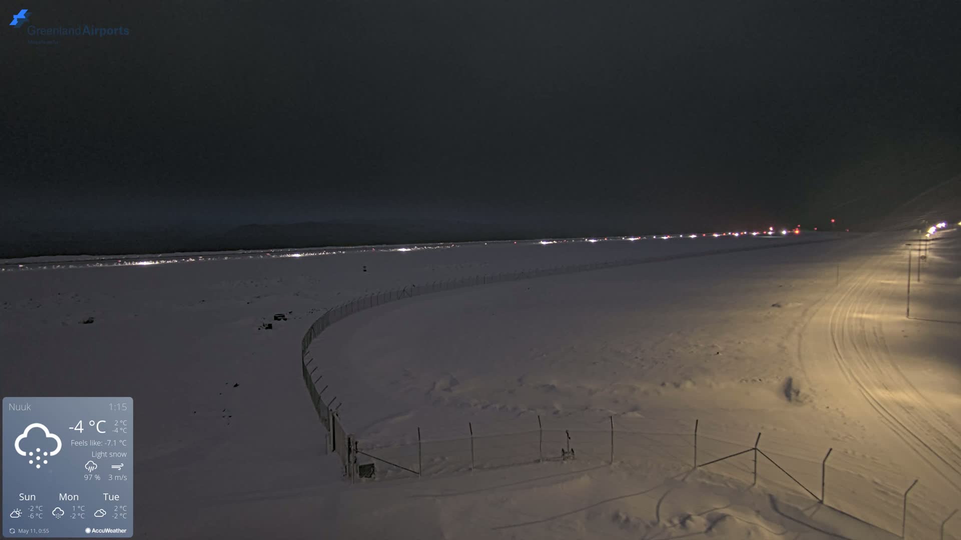 A snow-covered airfield at night under light snow, with a line of lights in the distance and a fence in the foreground.