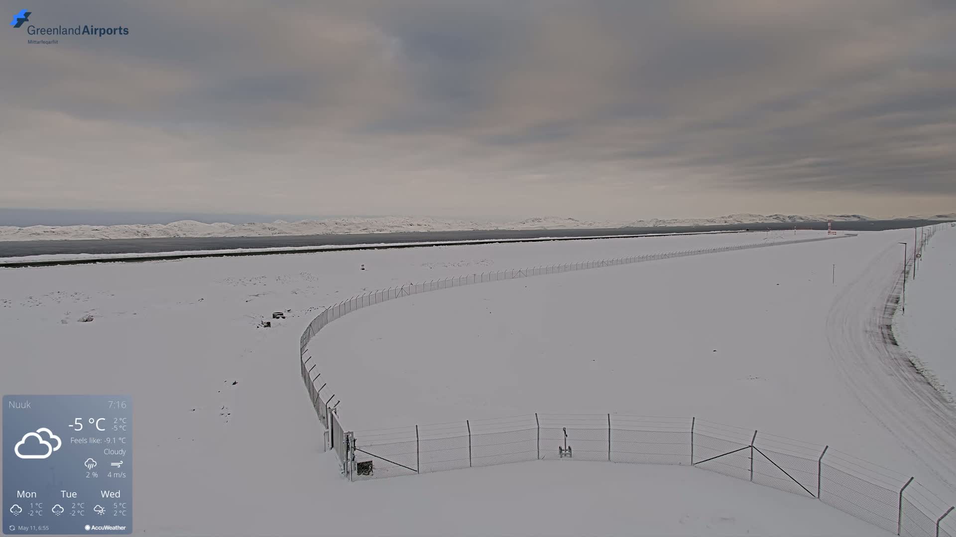 A snow-covered landscape with a chain-link fence curves across a wide, snow-covered expanse under a cloudy sky.