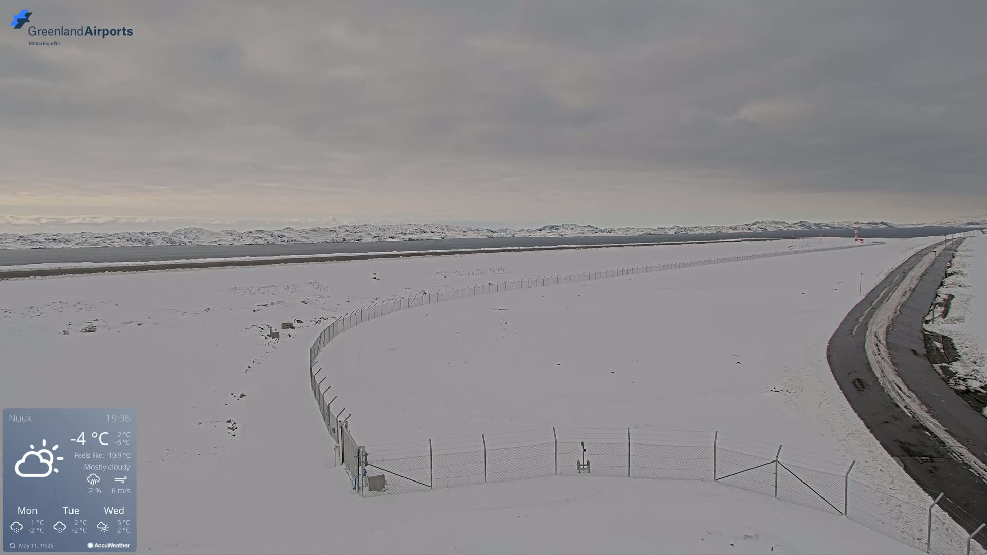A snow-covered airfield with a curving chain-link fence, a road, and a mostly cloudy sky in the background.