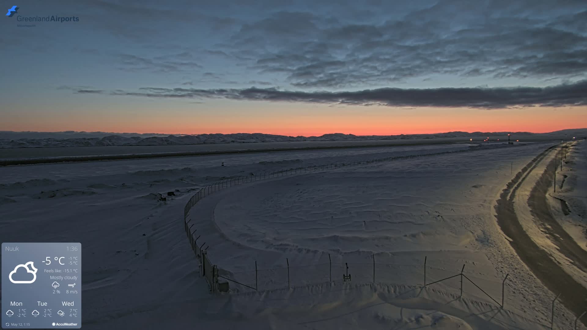 A snow-covered landscape at sunset under a mostly cloudy sky shows a fenced area near a road leading to a distant mountain range.
