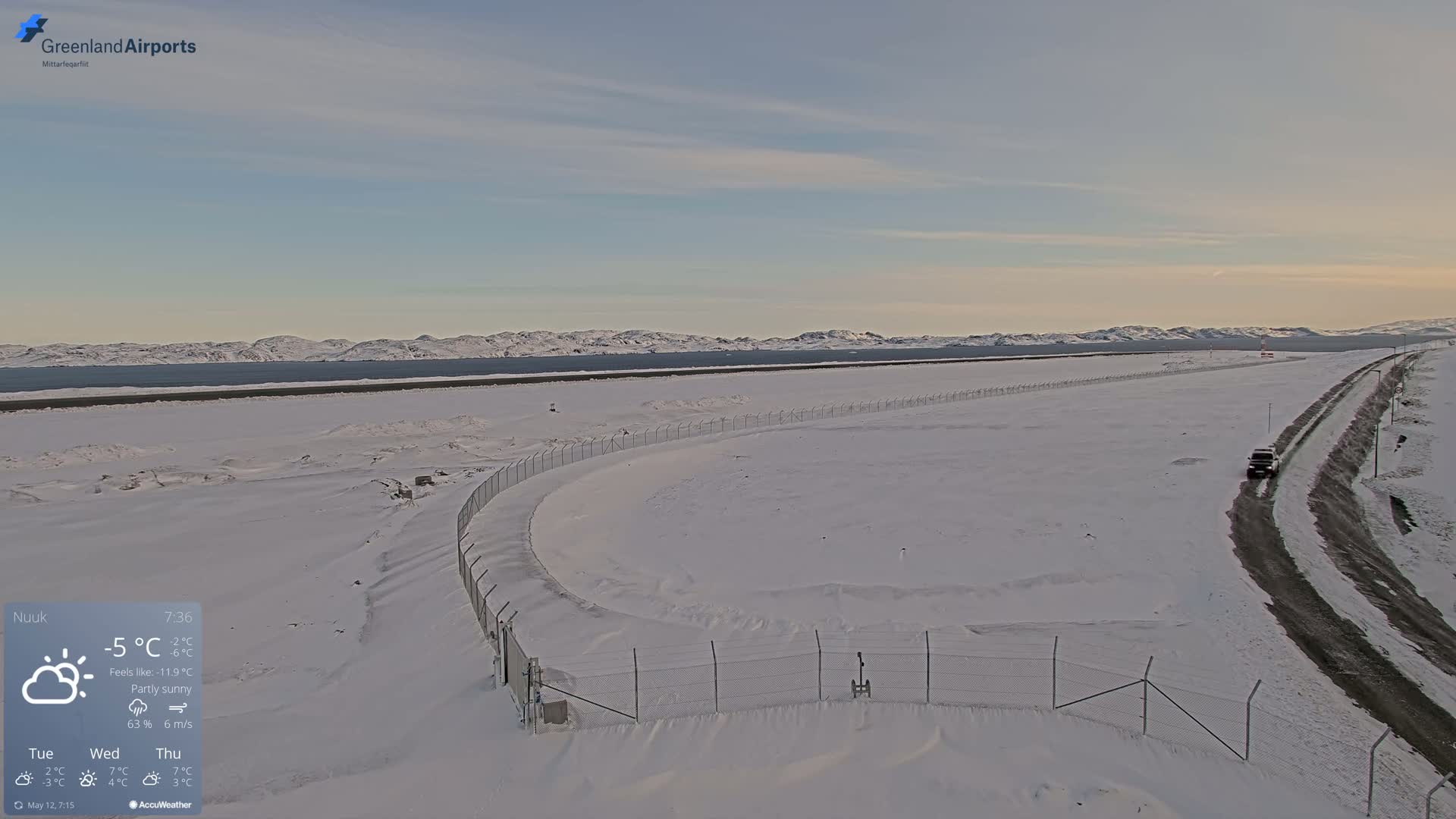 A snow-covered airport tarmac and road are visible under partly sunny skies, with mountains and a body of water in the background.