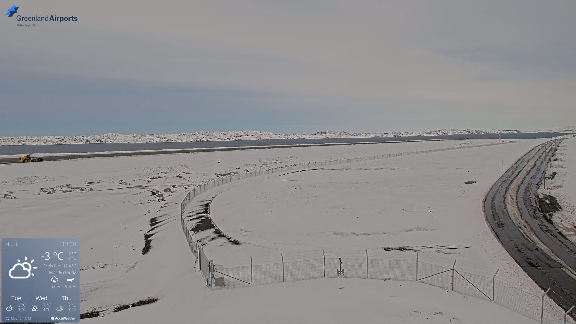 A snow-covered airport tarmac and road stretch toward a body of water under a mostly cloudy sky.