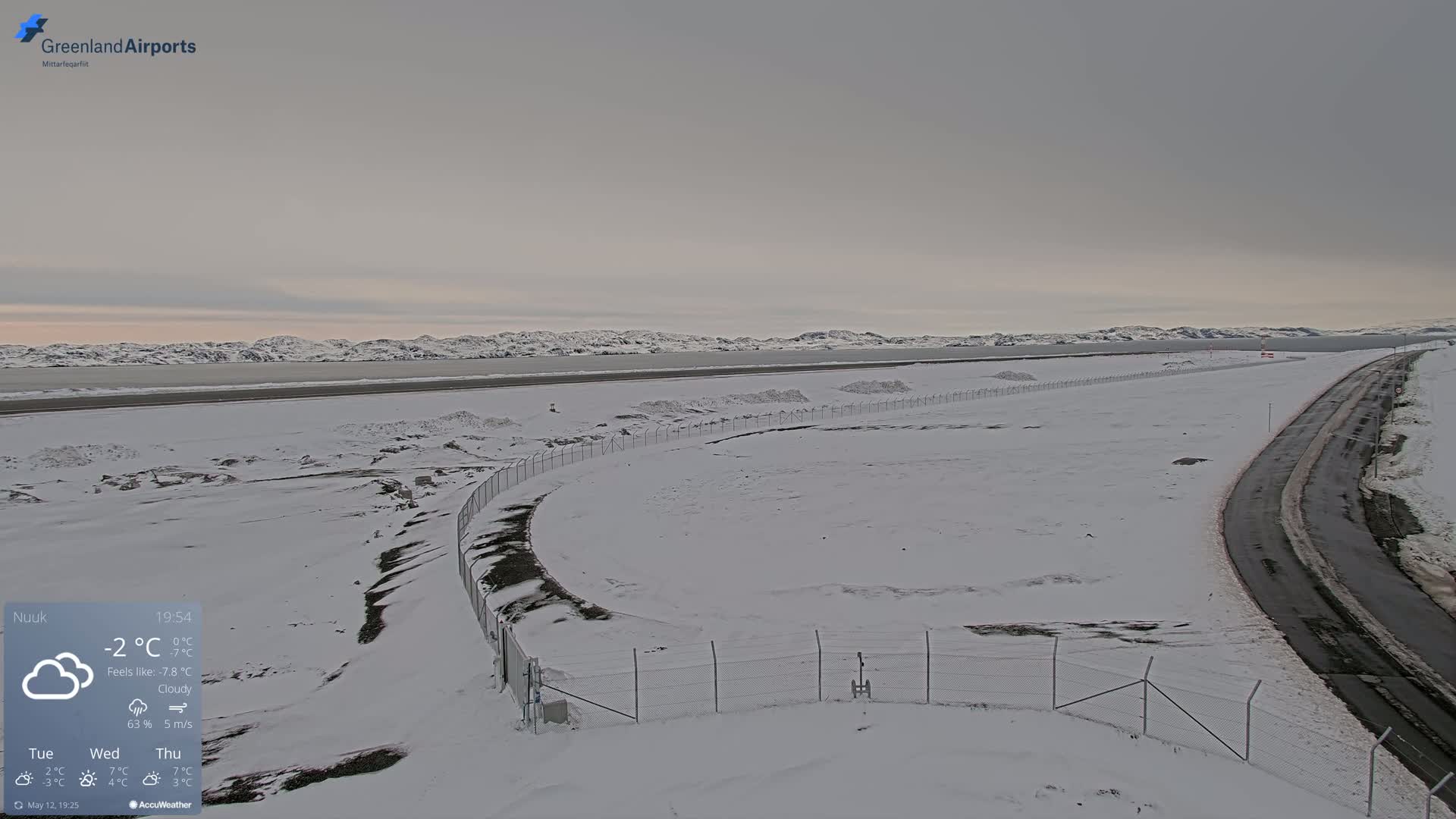 A snow-covered landscape with a road and a fenced-in area, overlooking a body of water and distant mountains under cloudy skies.