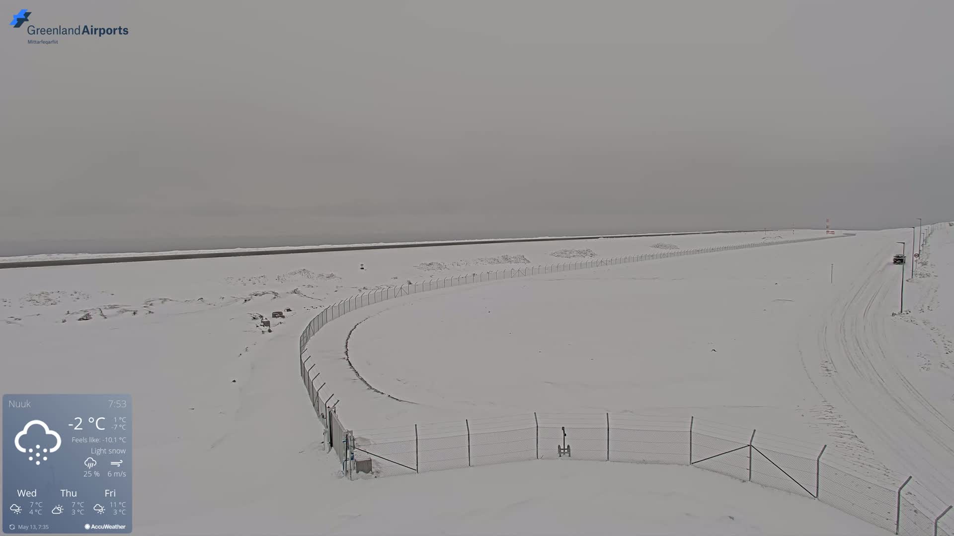 A snow-covered landscape with a chain-link fence curves around a section of a road, under a light snow.