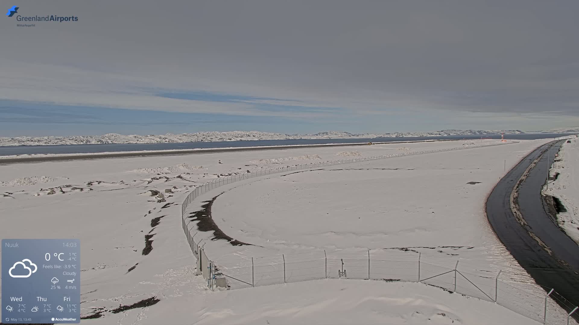 A snow-covered airport runway and road extend toward a body of water and distant, snow-capped mountains under a cloudy sky.
