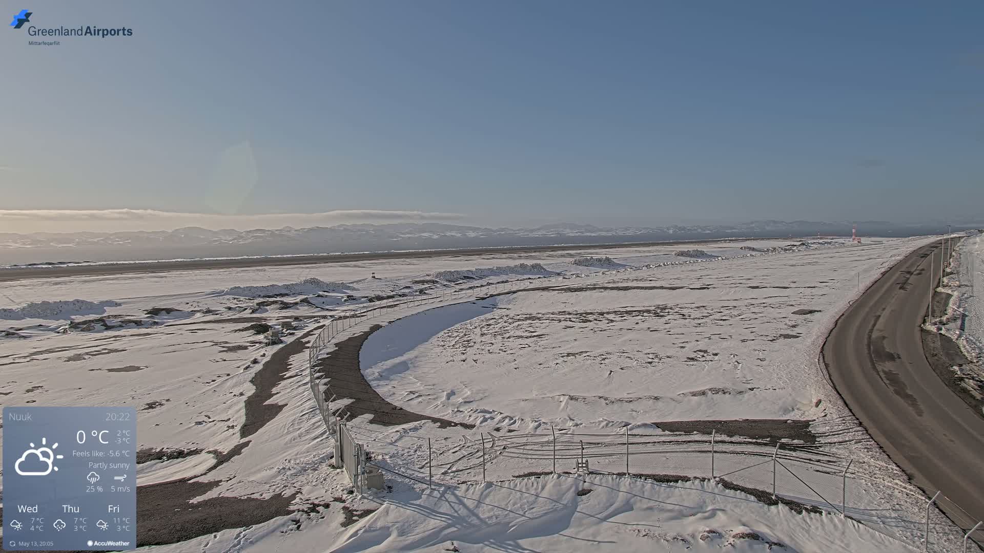 A snow-covered airfield with a road curving alongside a chain-link fence, under a partly sunny sky and distant mountains.