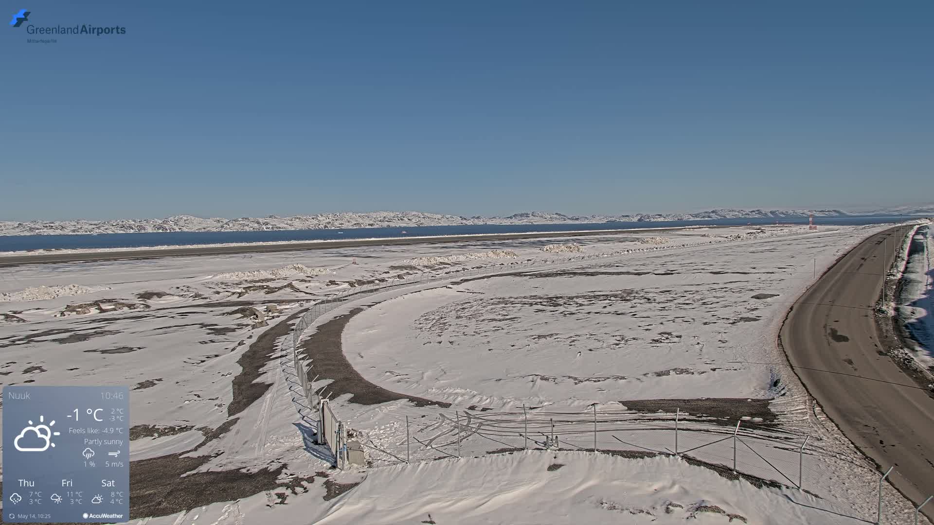 A snow-covered airport and road are visible, extending to a partly sunny, distant shoreline with snow-capped mountains under a clear blue sky.