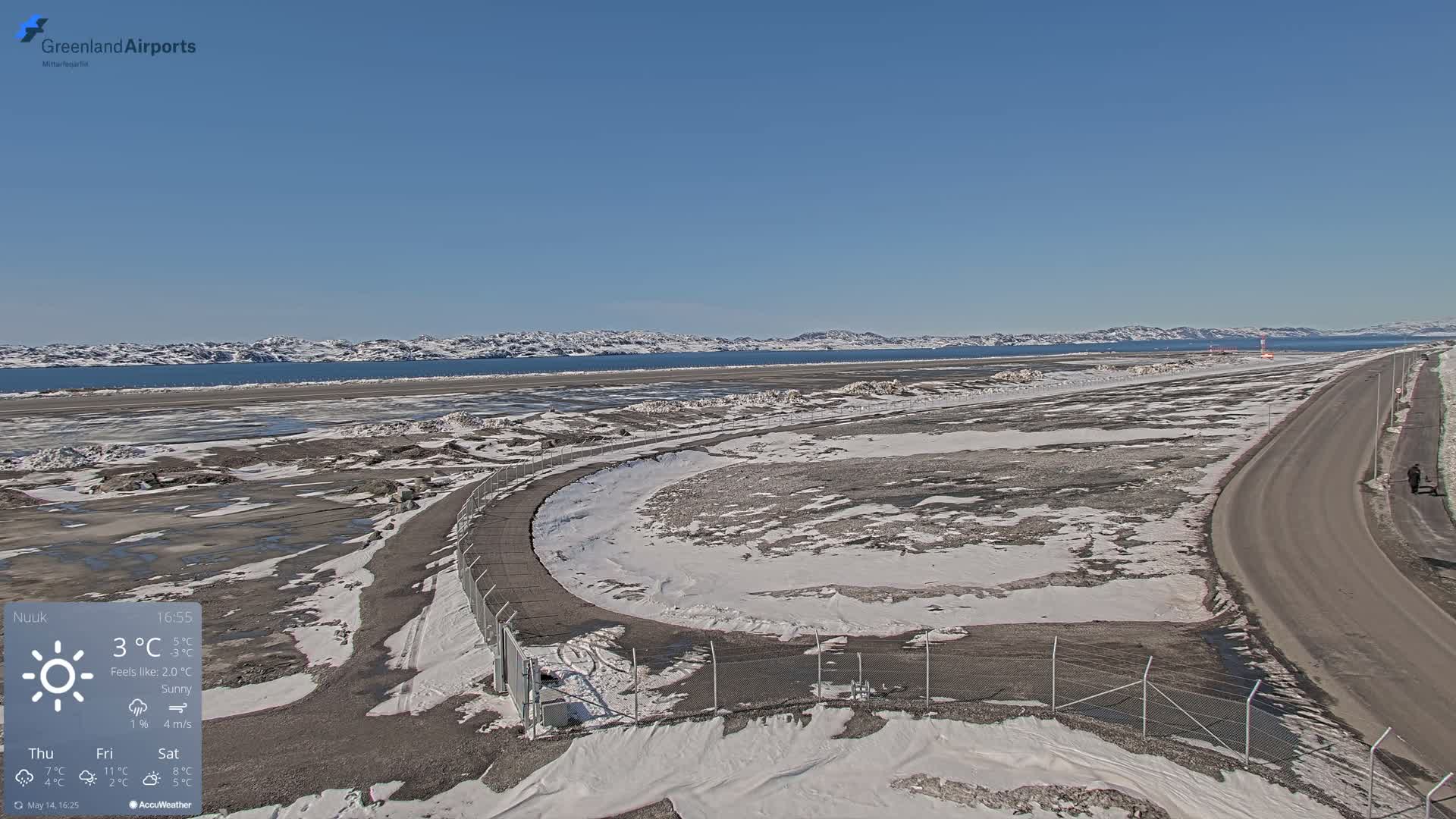 A sunny, snow-covered airport tarmac and roadway extend to a frozen body of water backed by distant mountains under a clear blue sky.