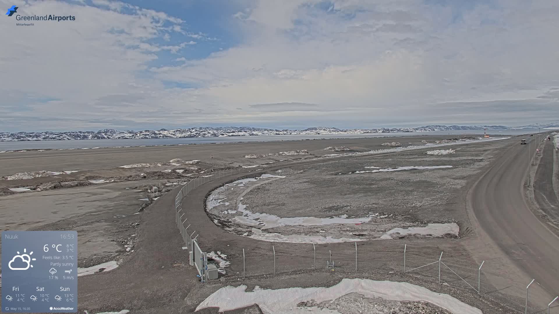 A partly sunny airfield with a road and snow patches on the ground, overlooked by a snow-capped mountain range in the distance.