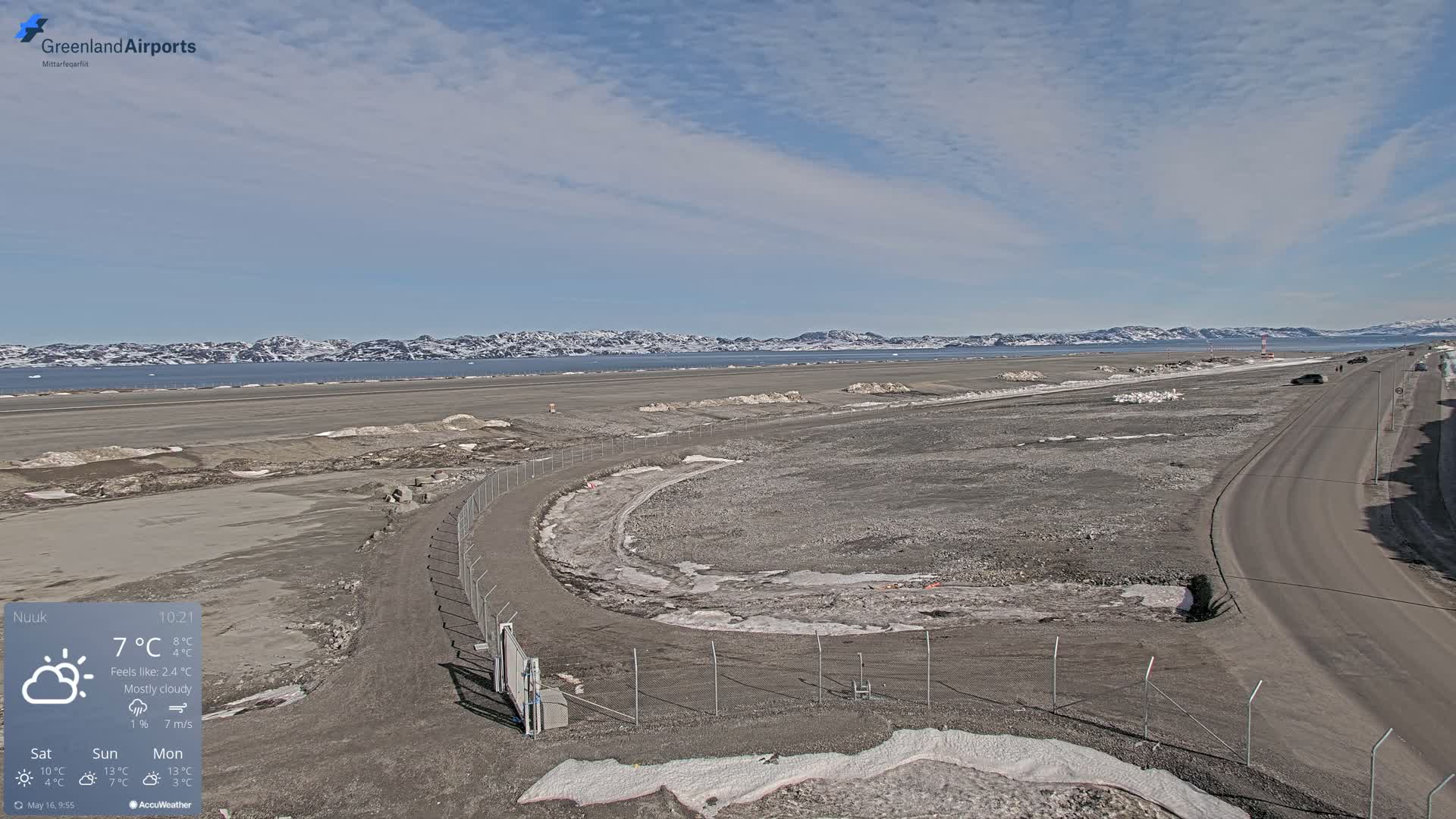 A mostly cloudy day reveals a flat, grayish-brown landscape with a road and a chain-link fence near a body of water with distant snow-capped mountains under a light blue sky.