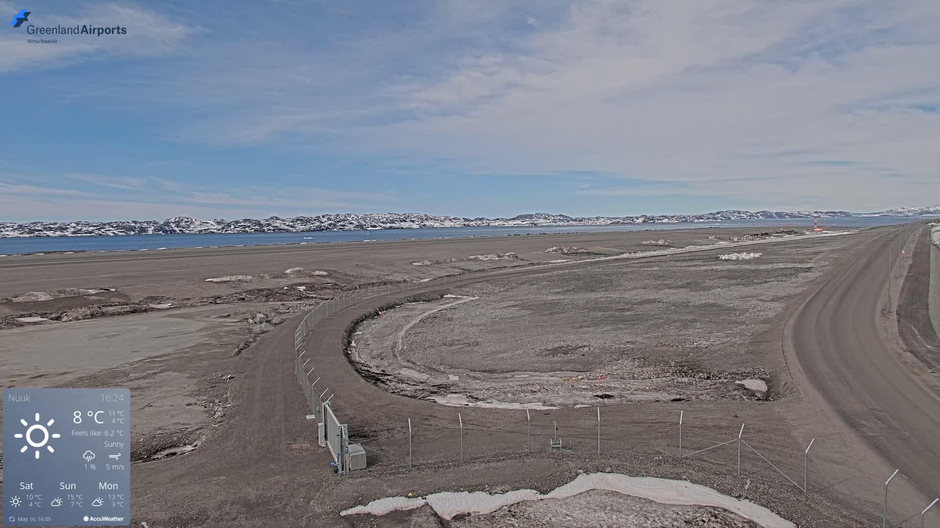 A sunny, sparsely vegetated airfield with a body of water and snow-capped mountains in the distance.