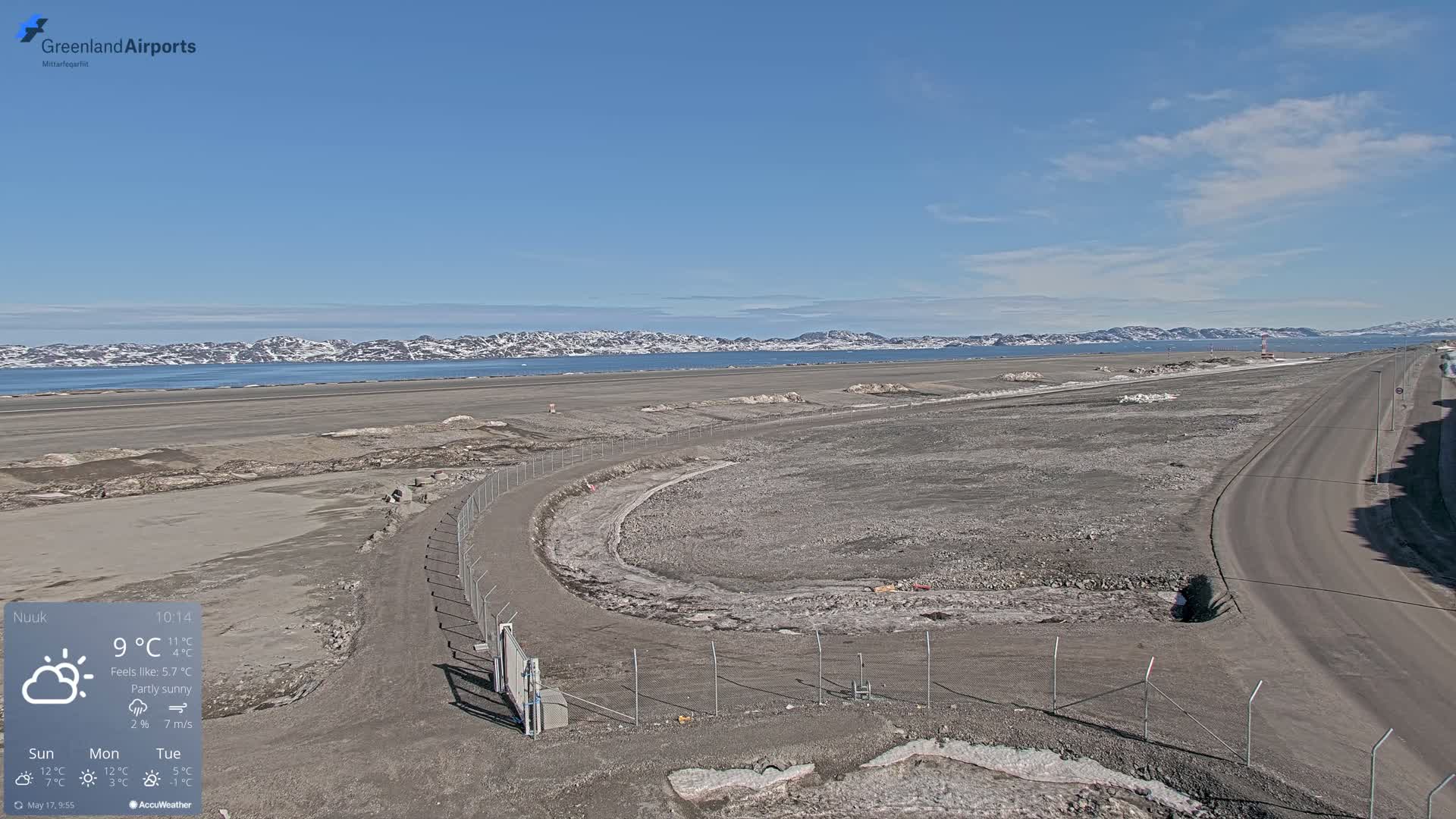 A mostly sunny, partly cloudy day reveals a wide, flat expanse of land next to a body of water with snow-covered mountains in the distance.