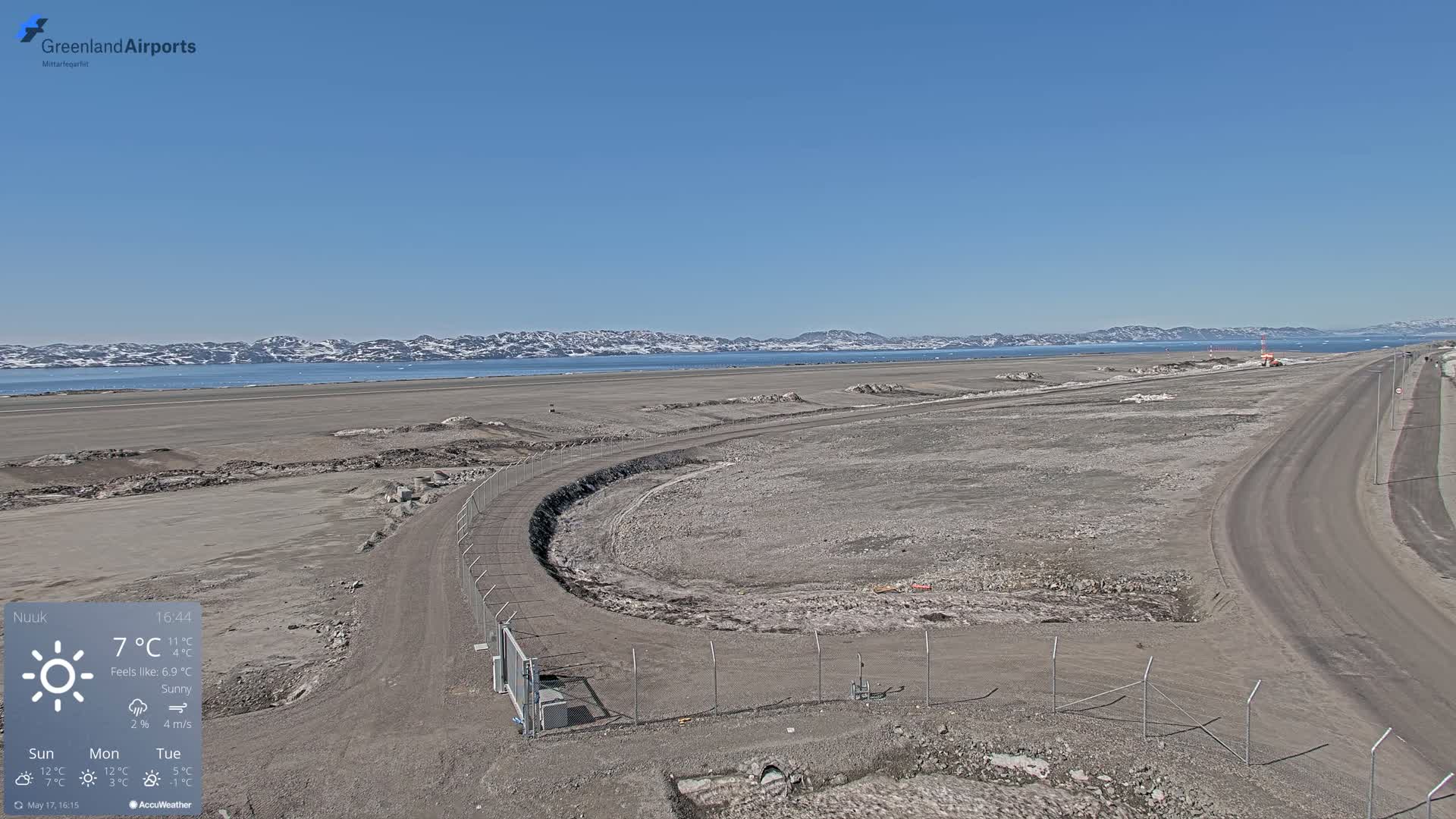 A sunny, sparsely vegetated airfield with a fenced perimeter and a road leading to it, overlooks a calm body of water and snow-capped mountains in the distance.
