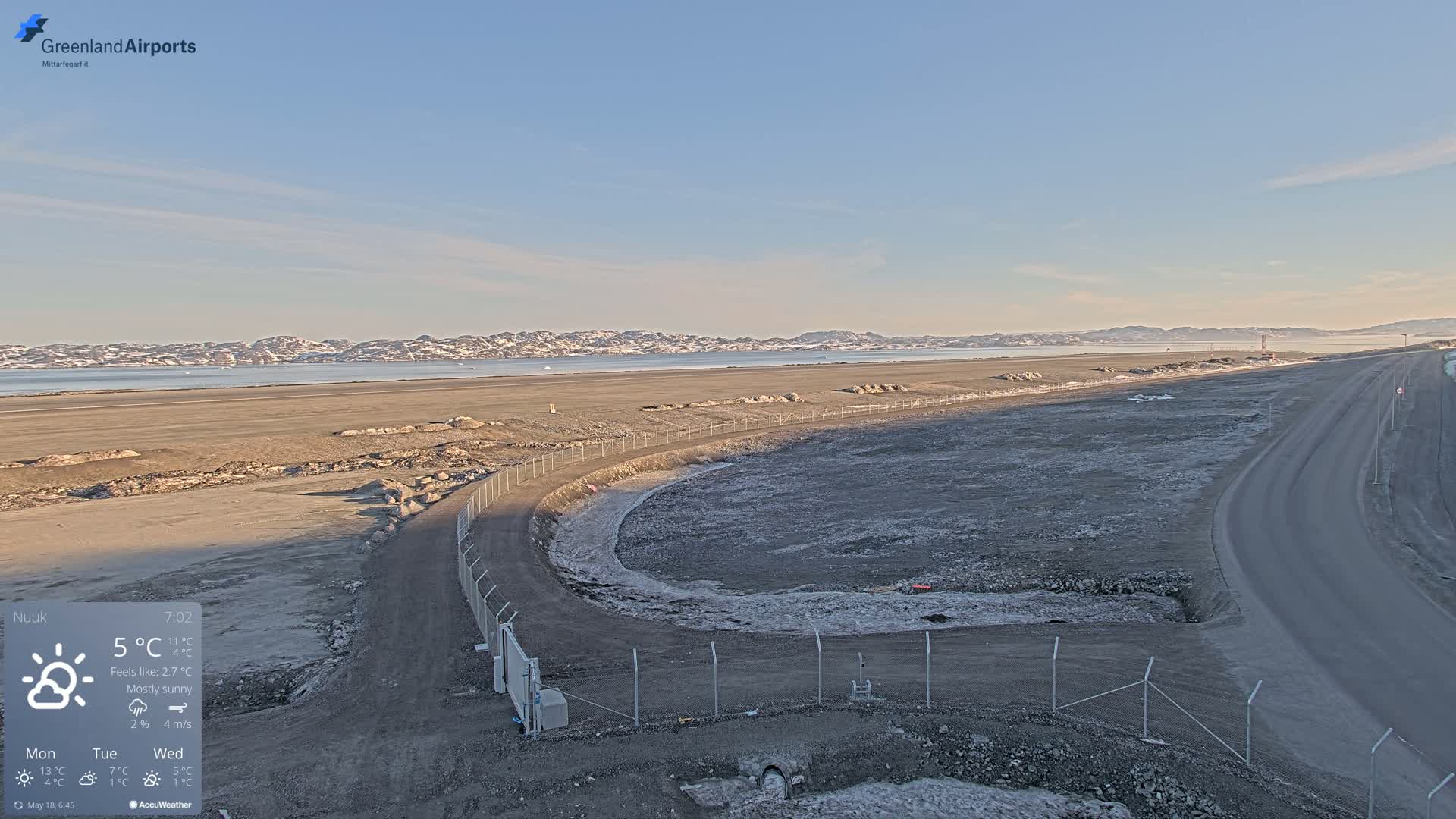 A mostly sunny, cool day reveals a wide expanse of flat, dark-colored land next to a body of water with distant snow-capped mountains under a clear pale blue sky.
