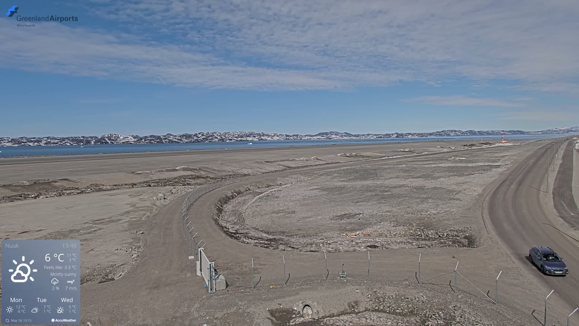 A mostly sunny, sparsely vegetated airfield with a road and a single car visible, overlooks a body of water with distant snow-capped mountains under a mostly clear sky.