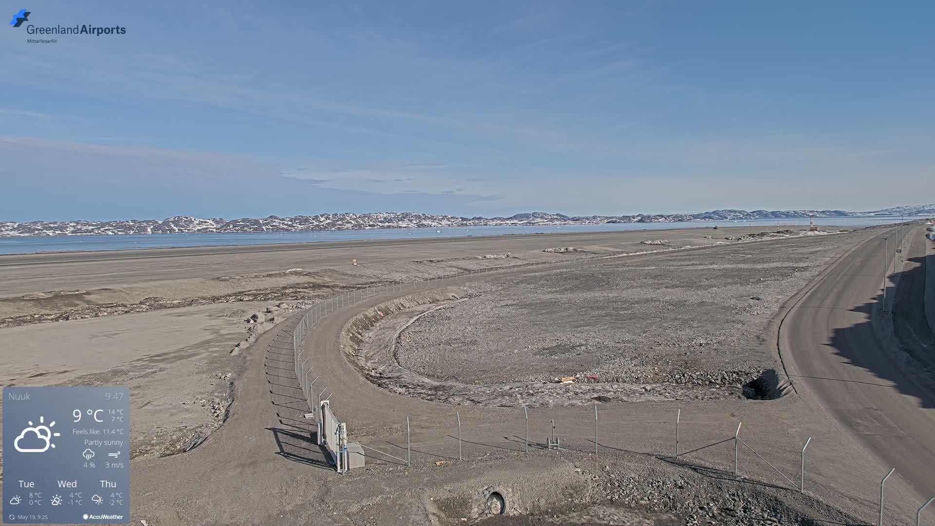A mostly sunny, partly cloudy day reveals a large, flat, undeveloped area of land adjacent to a paved road and a body of water with snow-capped mountains in the distance.