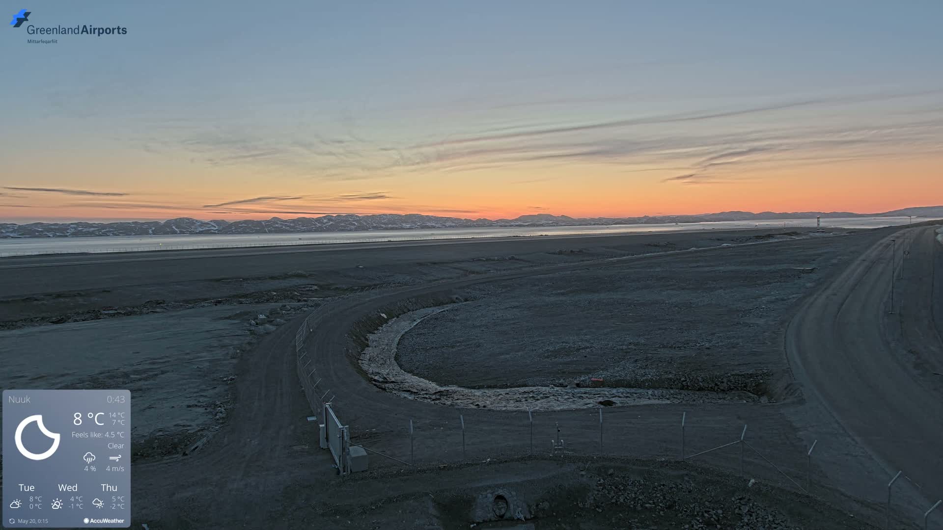 A wide, flat, dark-gray landscape with a curving road and a small stream under a clear, pinkish-orange sunset over distant mountains.