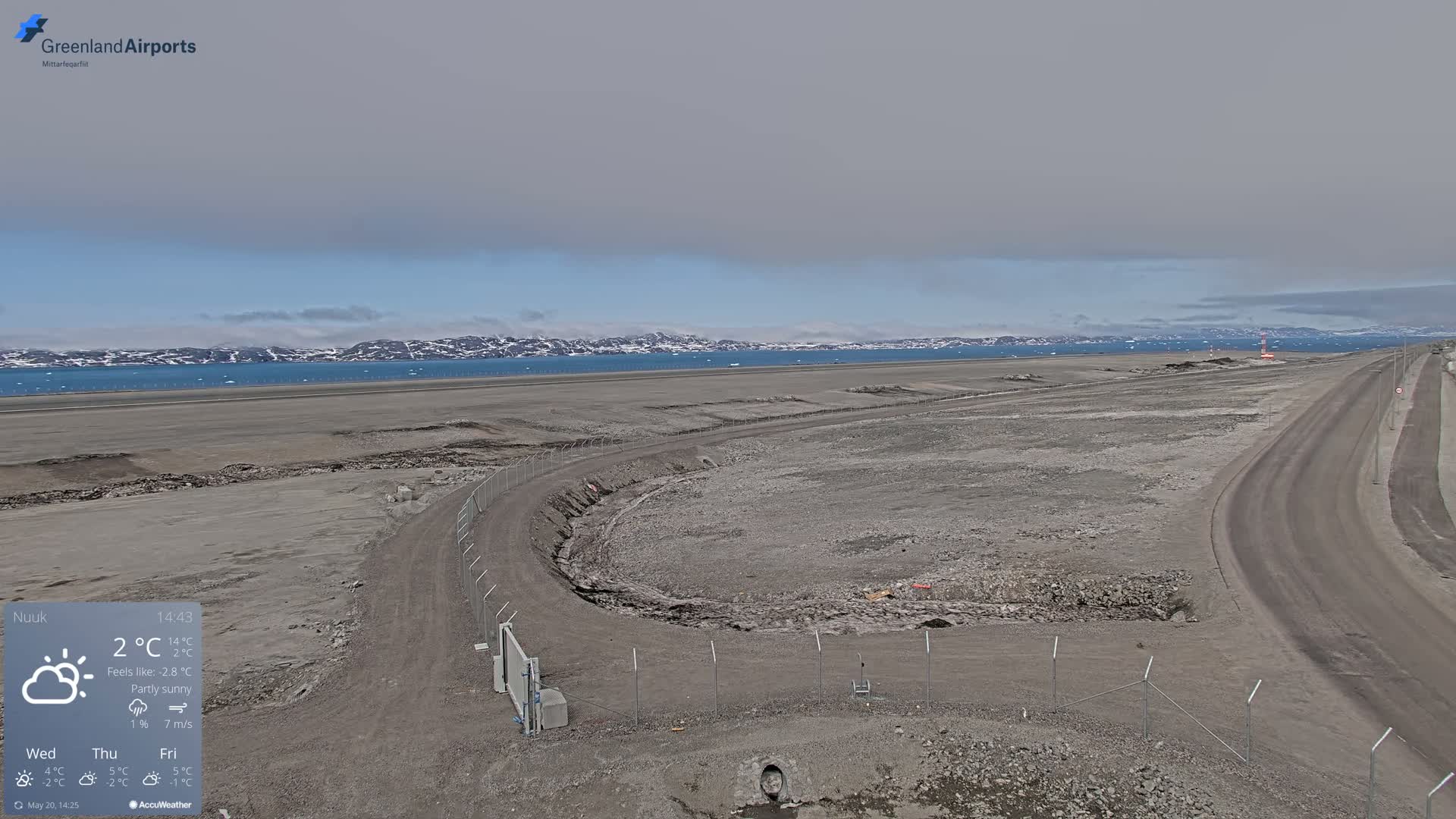 A mostly sunny, partly cloudy landscape shows a flat, gray, barren expanse of land with a road and a fence, overlooking a body of water with mountains in the distance.