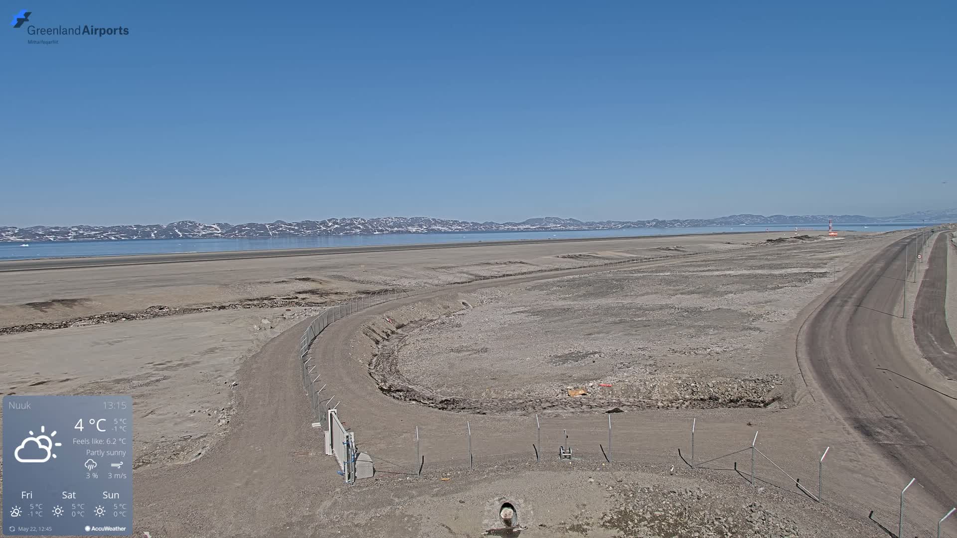 A partly sunny day reveals a flat, barren landscape with a body of water and distant snow-capped mountains in the background, featuring a roadway and what appears to be an airfield under construction.