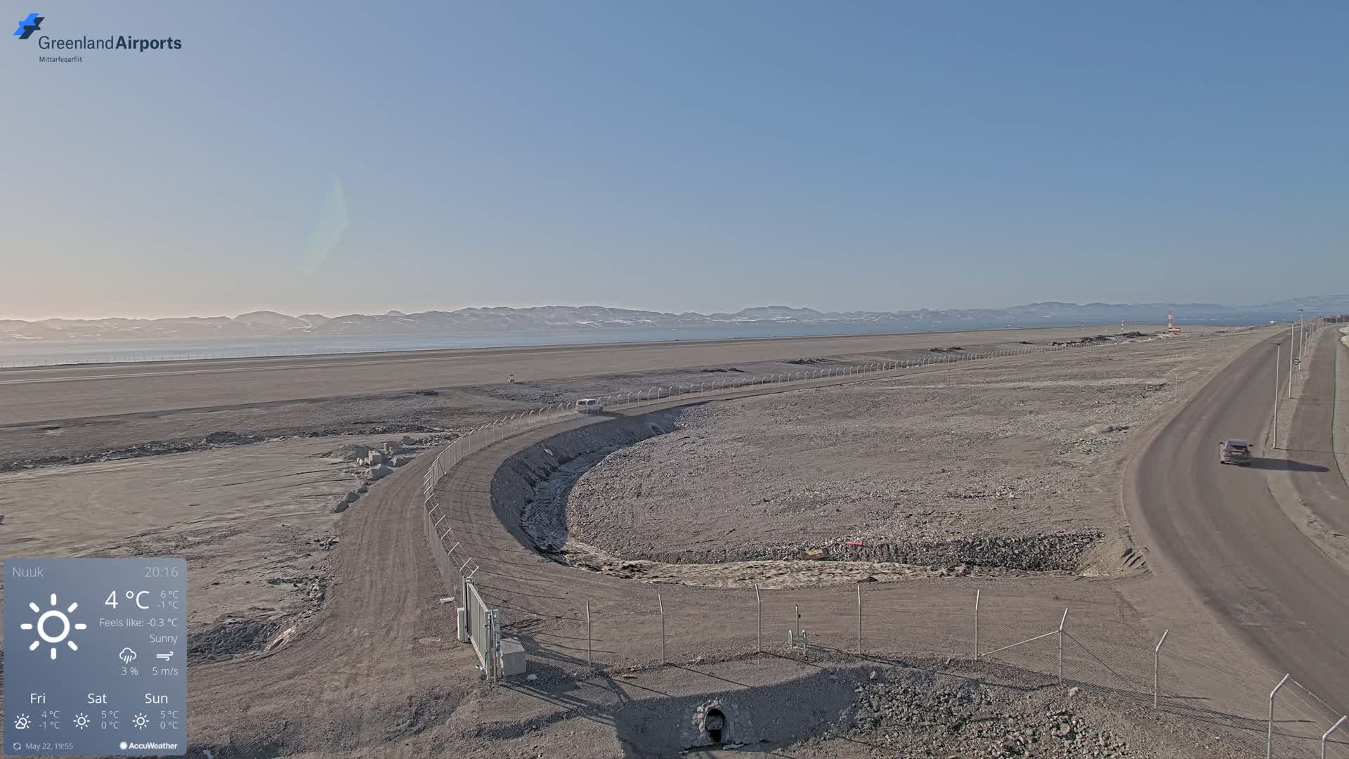 A sunny, flat, and barren landscape with a road and a vehicle, featuring a body of water and distant mountains in the background.