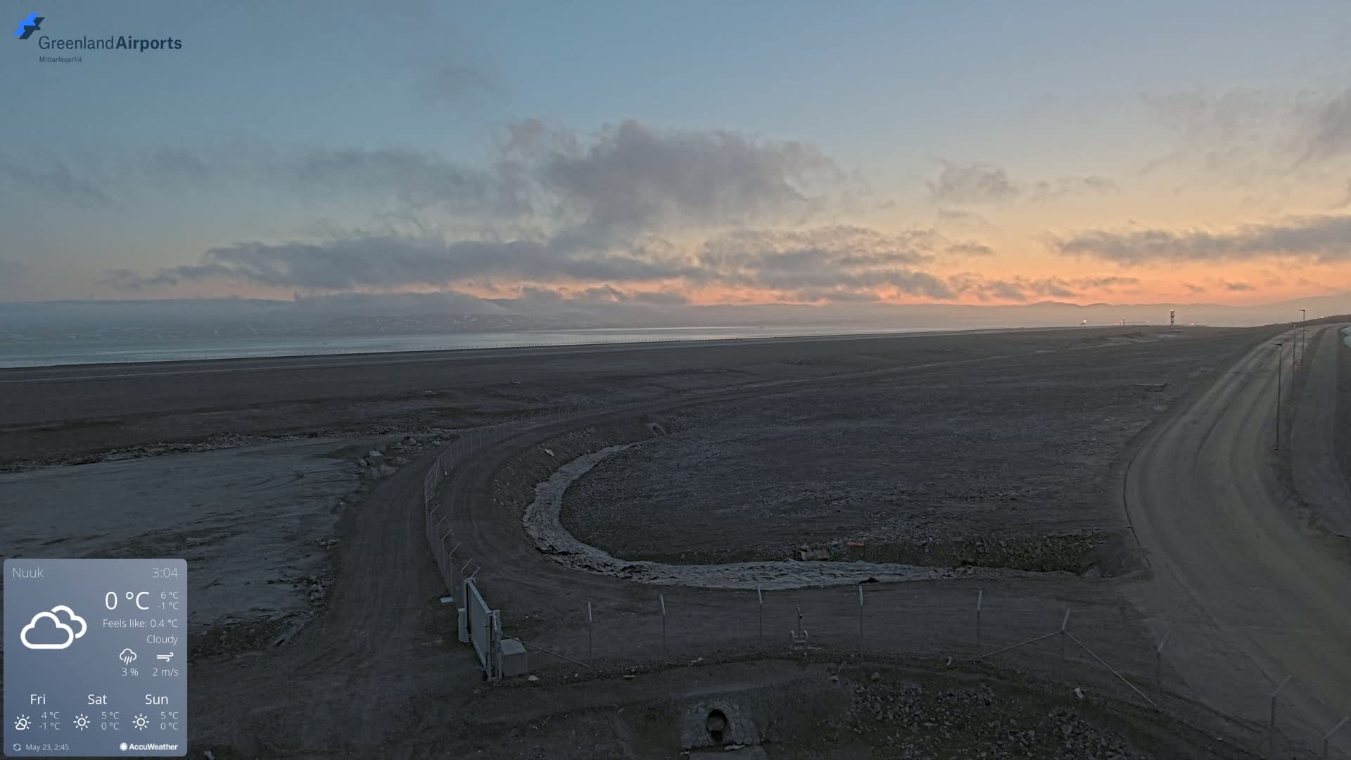 A cloudy sunset over a flat, dark landscape with a body of water in the distance and a curving road near a fenced area.