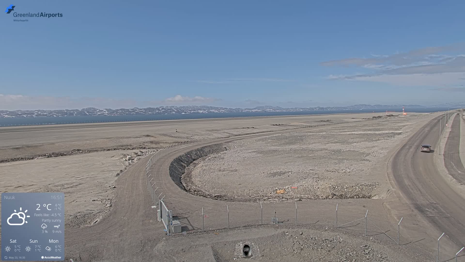 A mostly sunny, partly cloudy day shows an airport tarmac with a road curving around a fenced-in area under construction, mountains and water in the background.