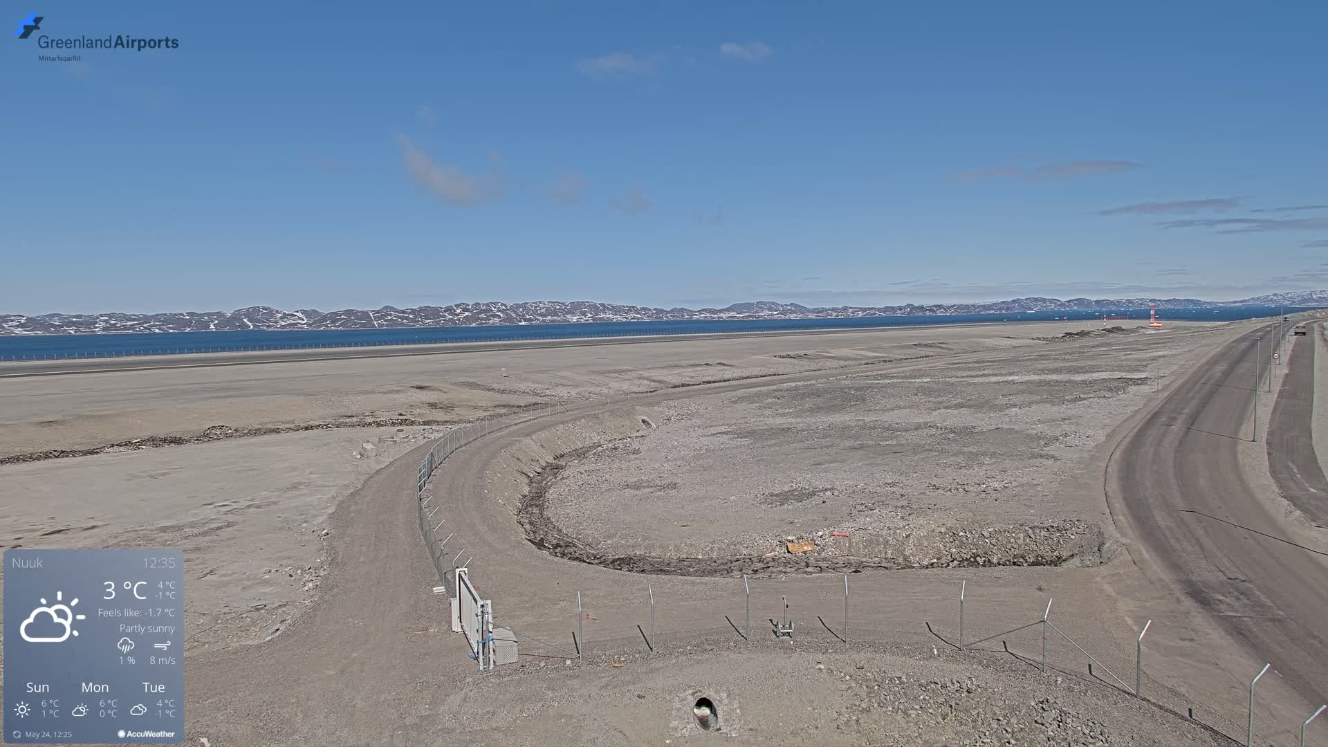 A partly sunny, 3°C day shows an airport tarmac under construction, next to a body of water with distant snow-capped mountains.