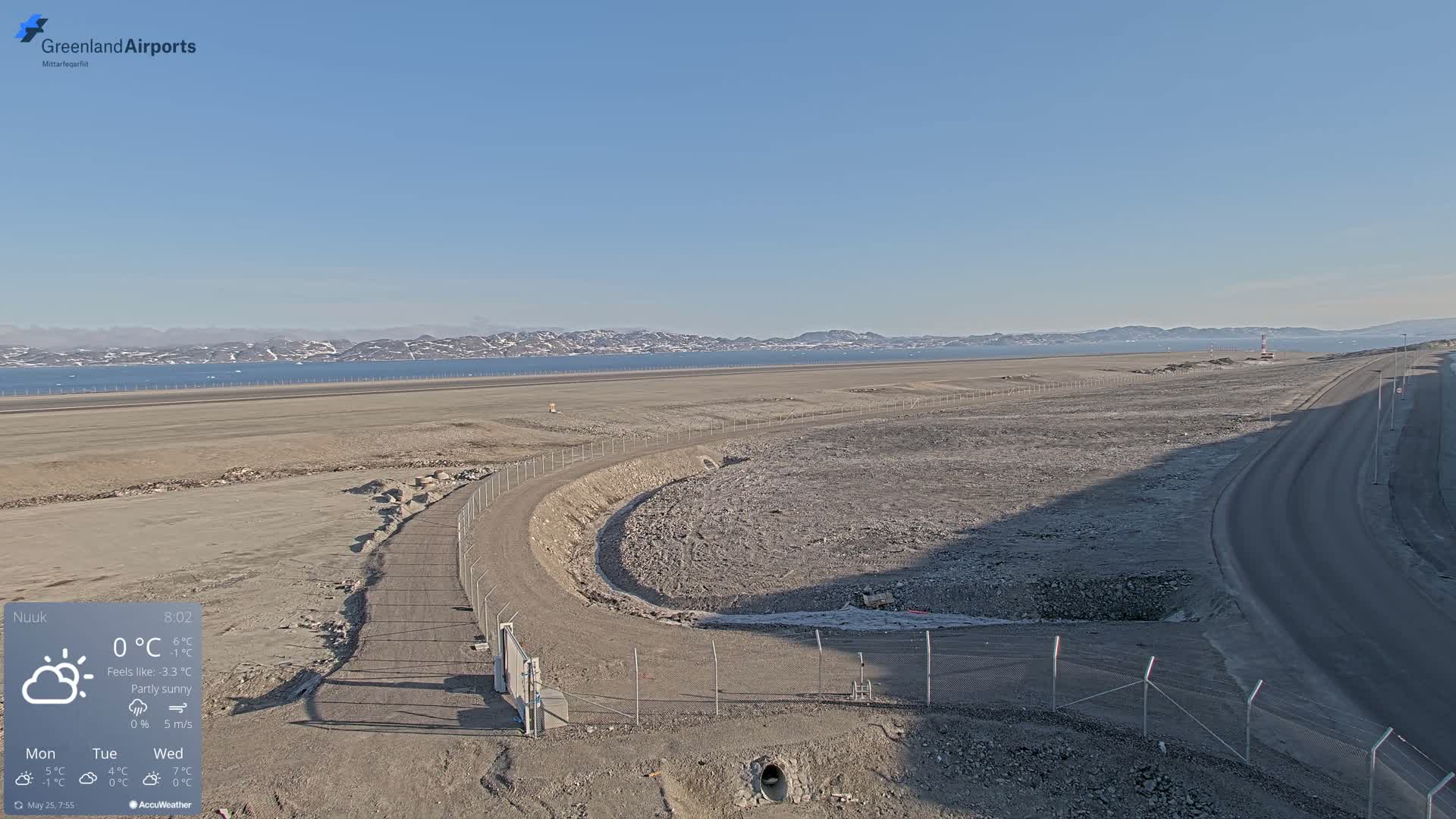 A mostly sunny, partly cloudy day shows a view of a sparsely vegetated, rocky airport area next to a body of water and distant mountains.