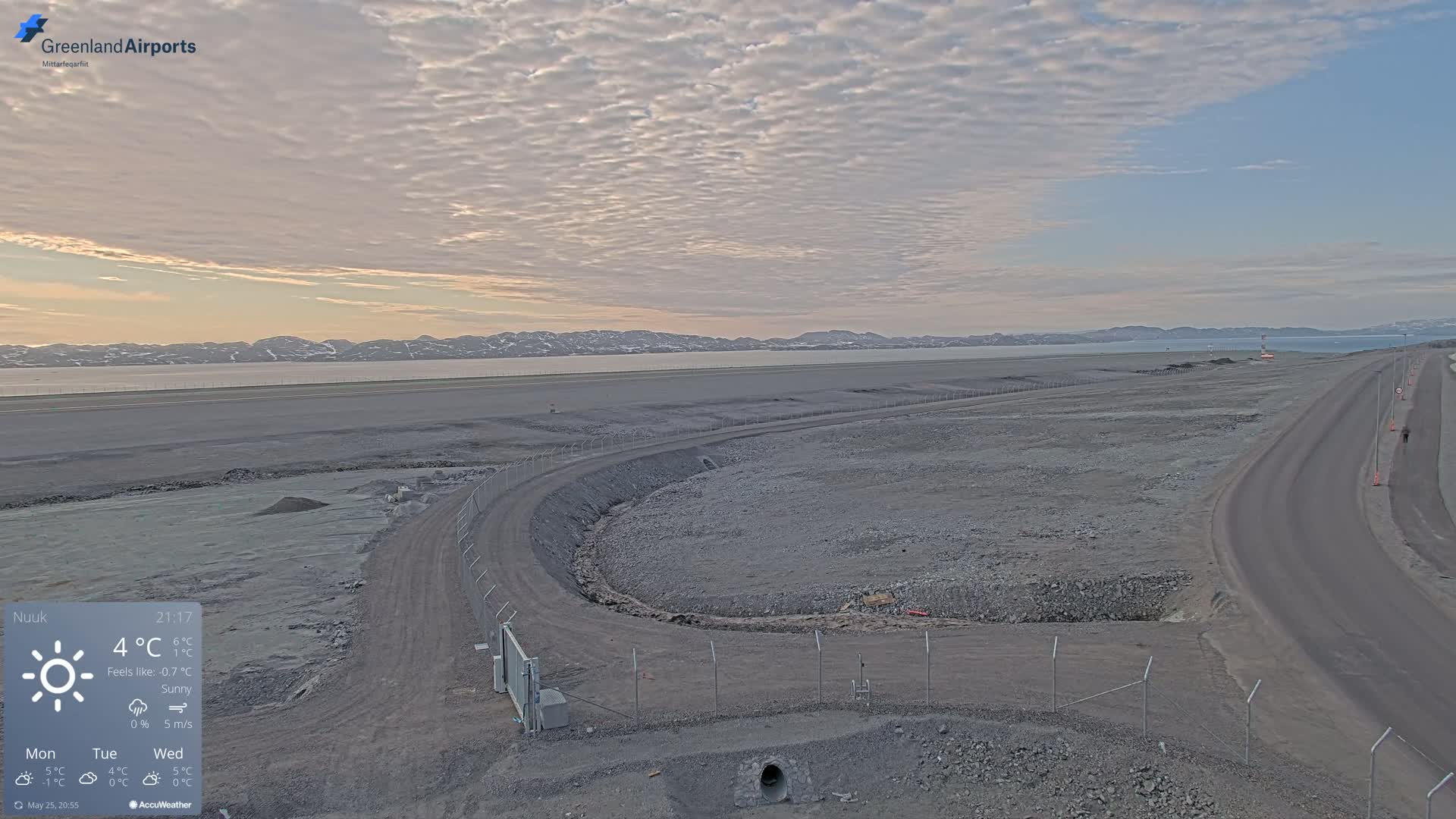 A mostly sunny, late afternoon view of a large, flat, rocky expanse with a road curving away from the foreground, a body of water in the distance, and mountains on the horizon.
