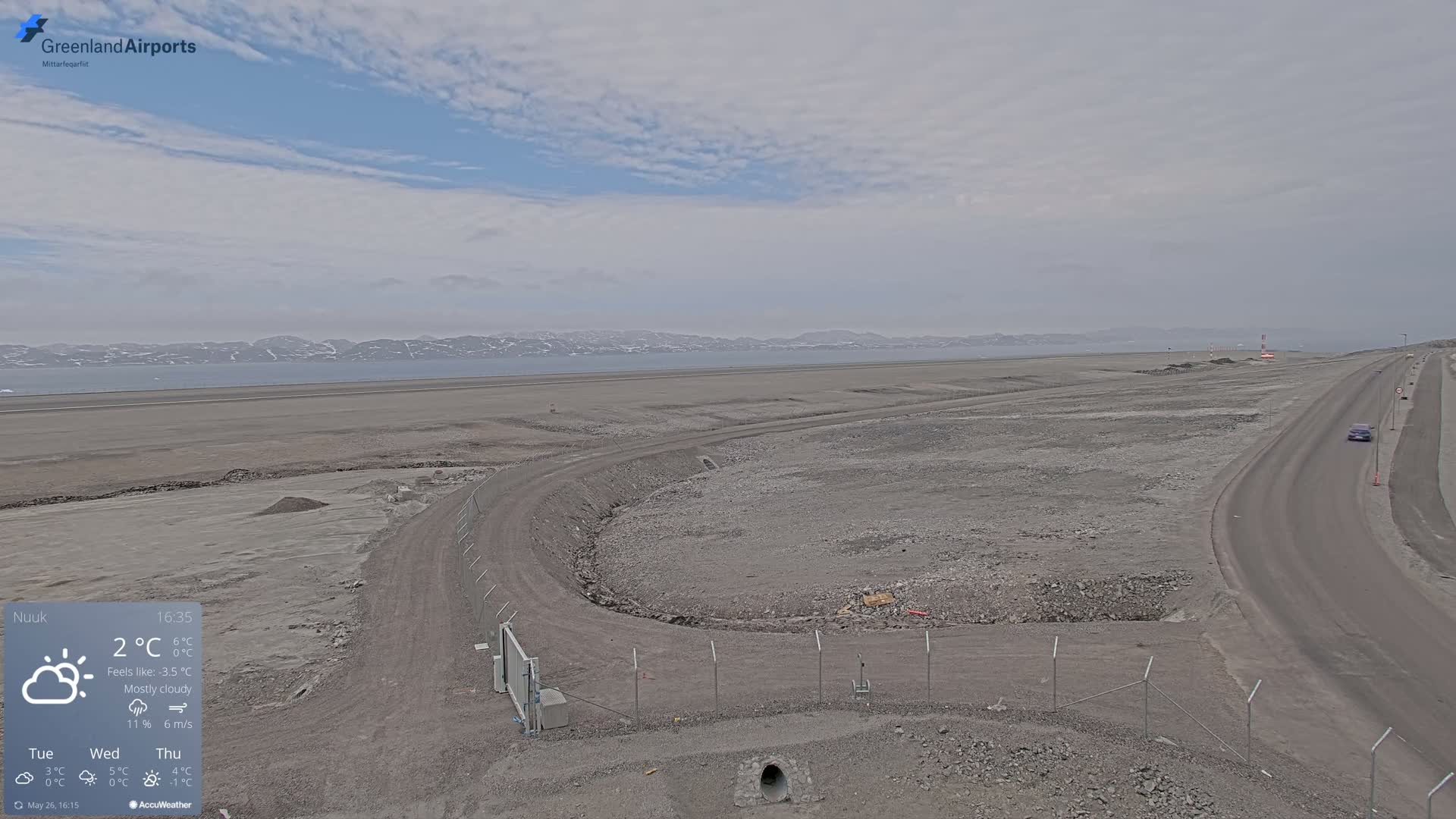A mostly cloudy day reveals a mostly barren landscape with a road and a fenced-in area under construction, next to a body of water and distant mountains.