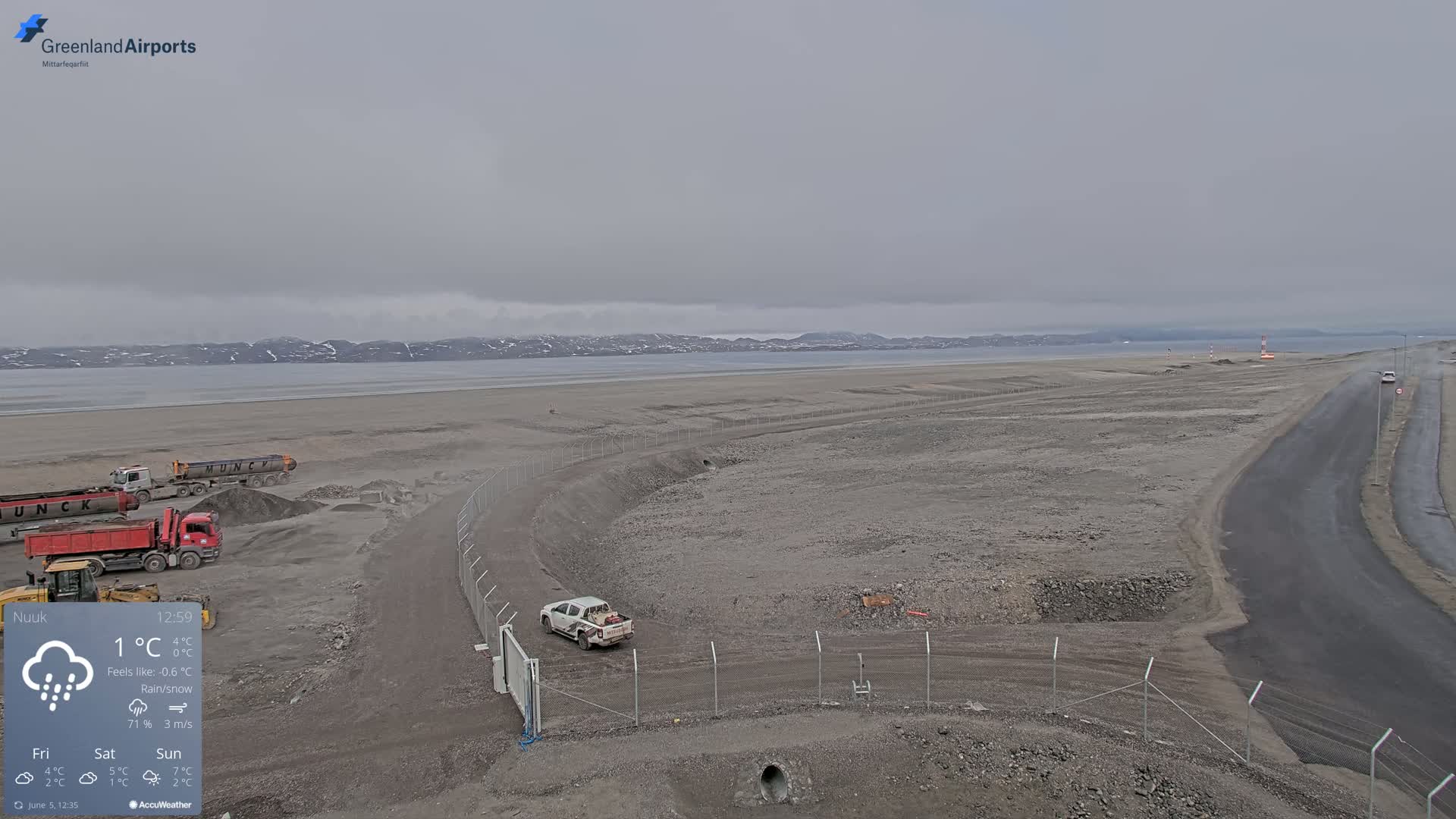 A flat, gray landscape with construction vehicles and a pickup truck, under a cloudy sky with light rain or snow.