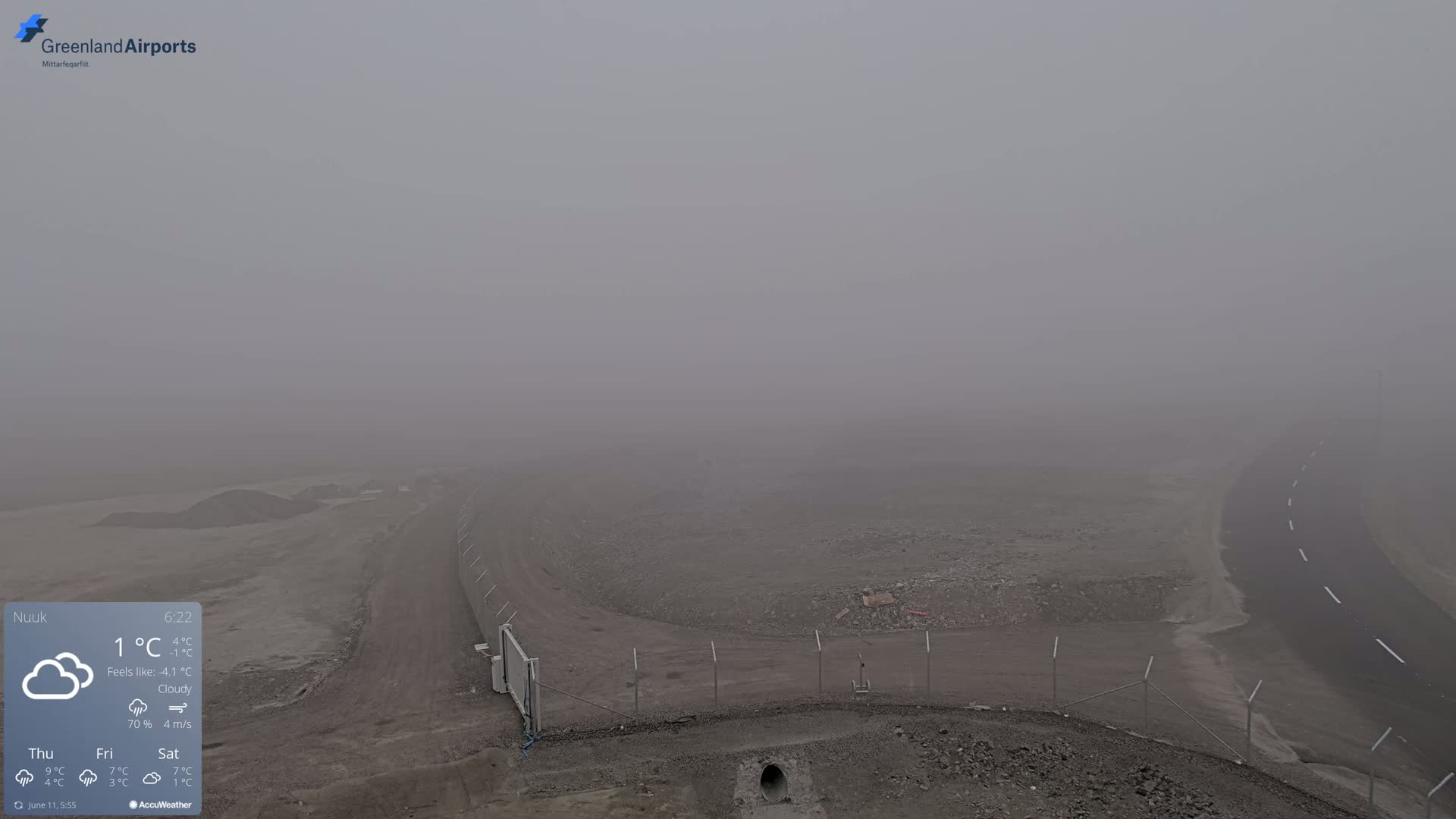 A foggy, cloudy outdoor scene shows a sparsely vegetated, rocky area with a paved road curving away into the thick fog.